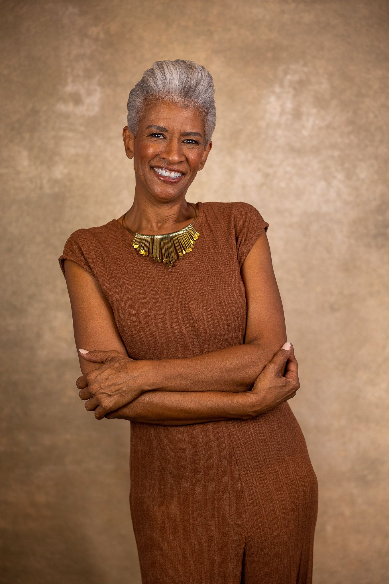 Professional portrait of woman in brown dress with statement necklace against textured bronze background.