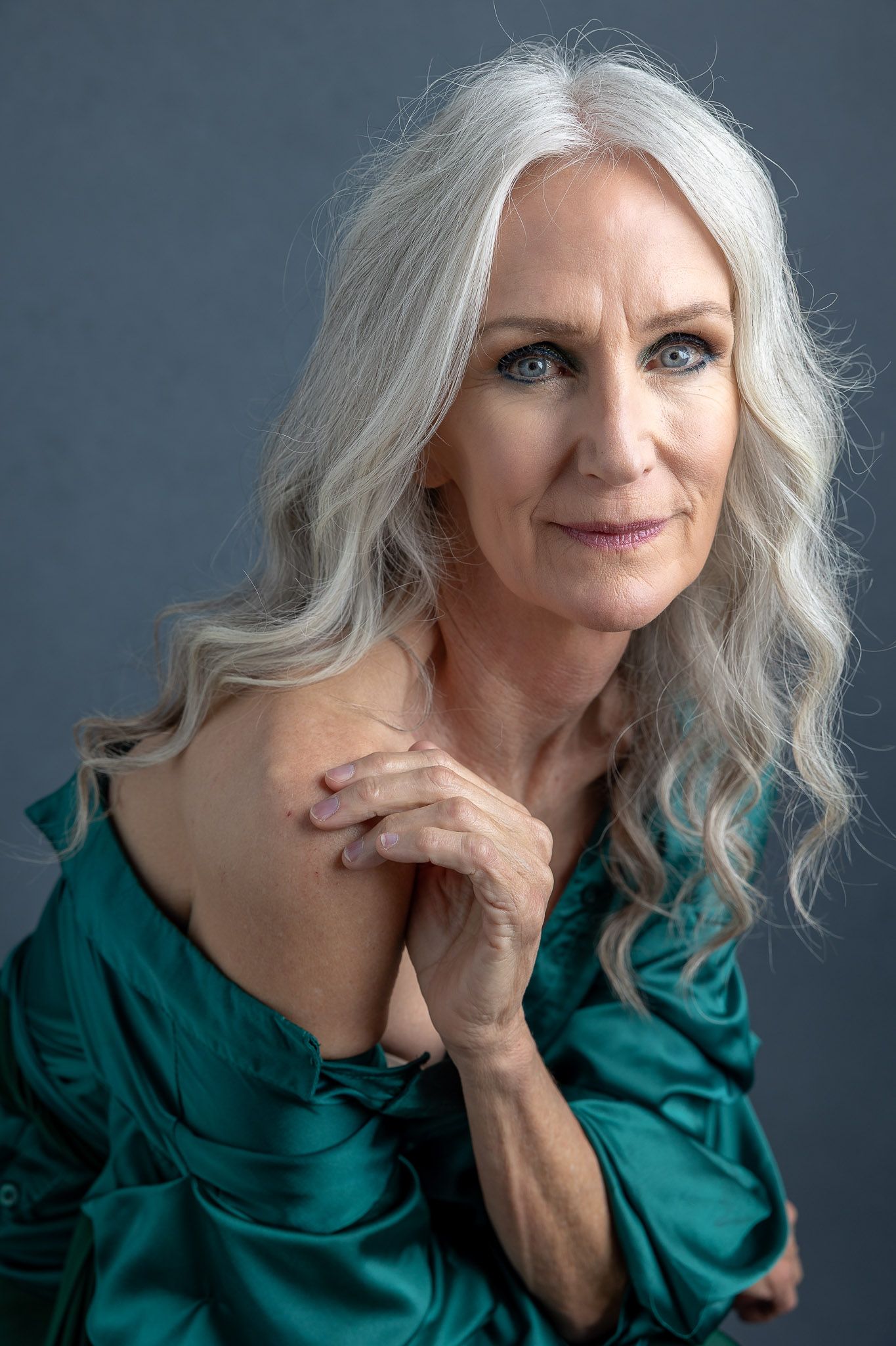 Elegant portrait of woman with long silver hair wearing emerald green silk blouse against grey background.