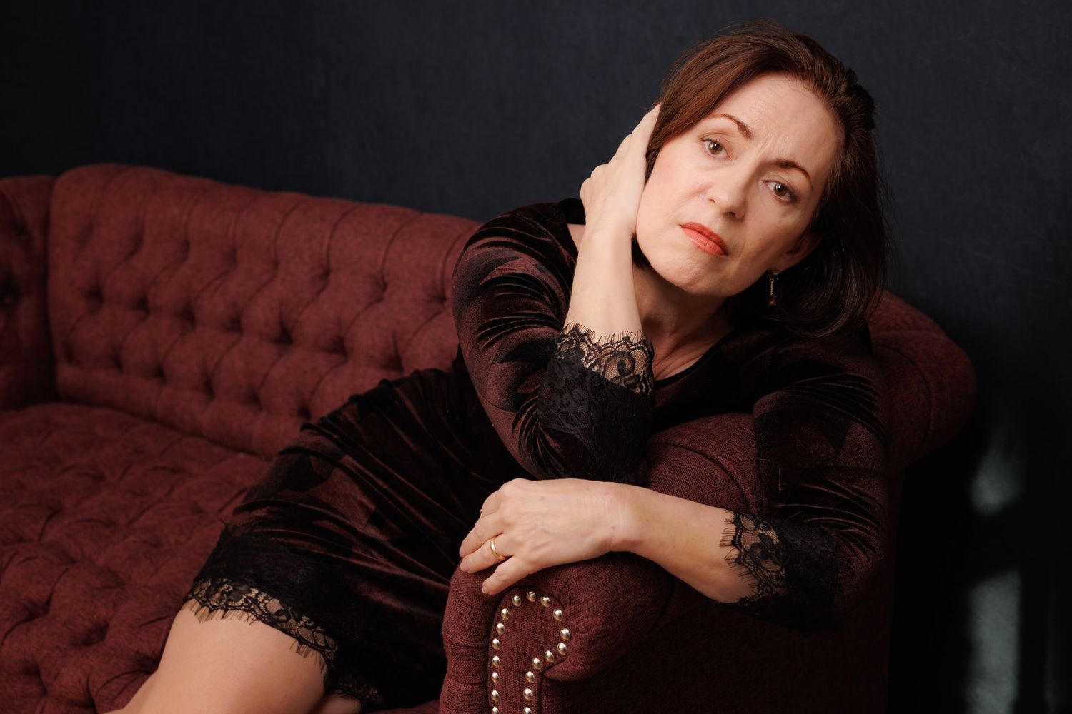Portrait of a model in vintage-style velvet dress reclining on burgundy tufted couch.