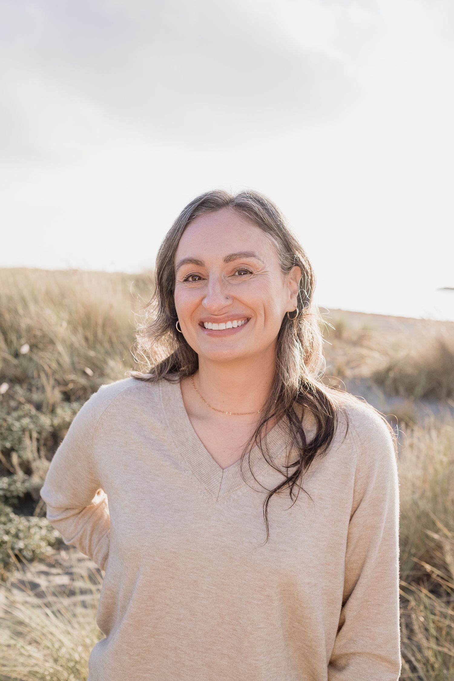 image of Angela Tellez, professional photographer at the beach. Dunes and beach grass. Woman with dark hair and smiling at the camera.