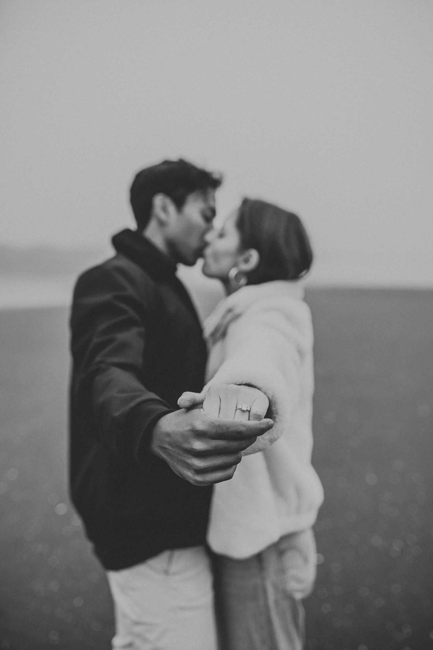 close up of engagement ring in the foreground while couple kisses in background