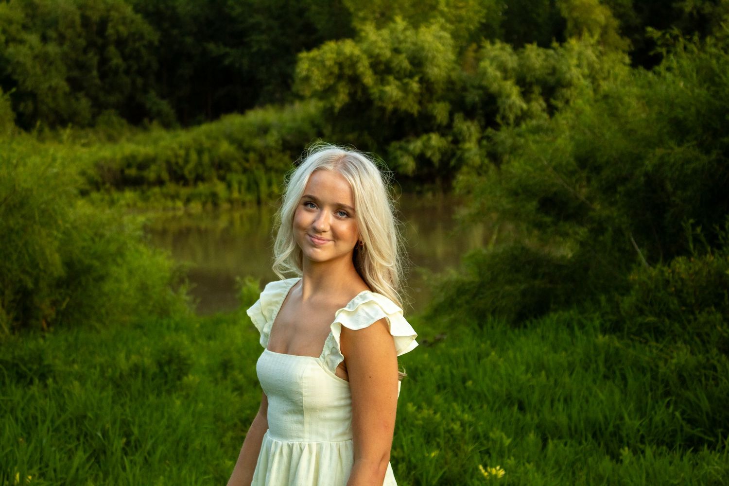 Woman in white dress poses outdoors in natural green setting during golden hour.
