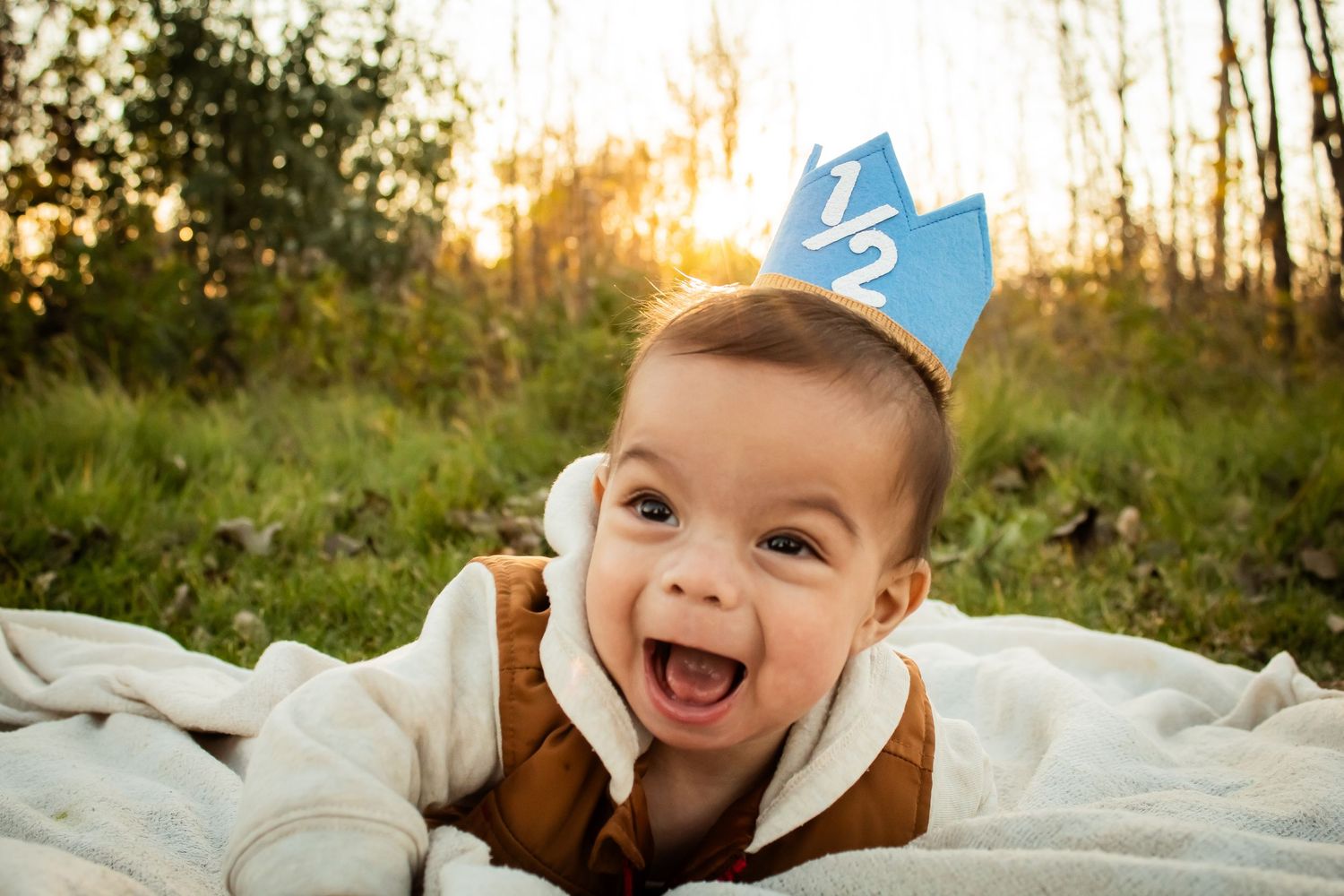 Baby wearing blue birthday hat smiling on white blanket outdoors.