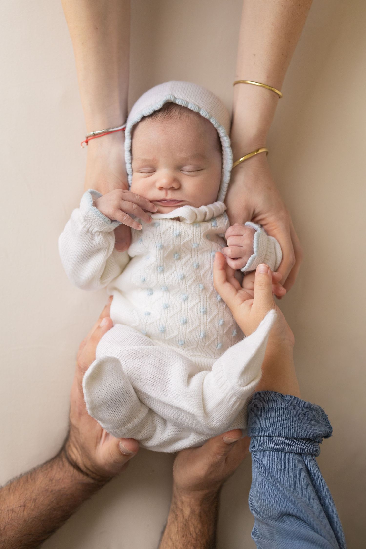 Newborn baby in white onesie and bonnet held gently by parents hands during professional newborn photography session.