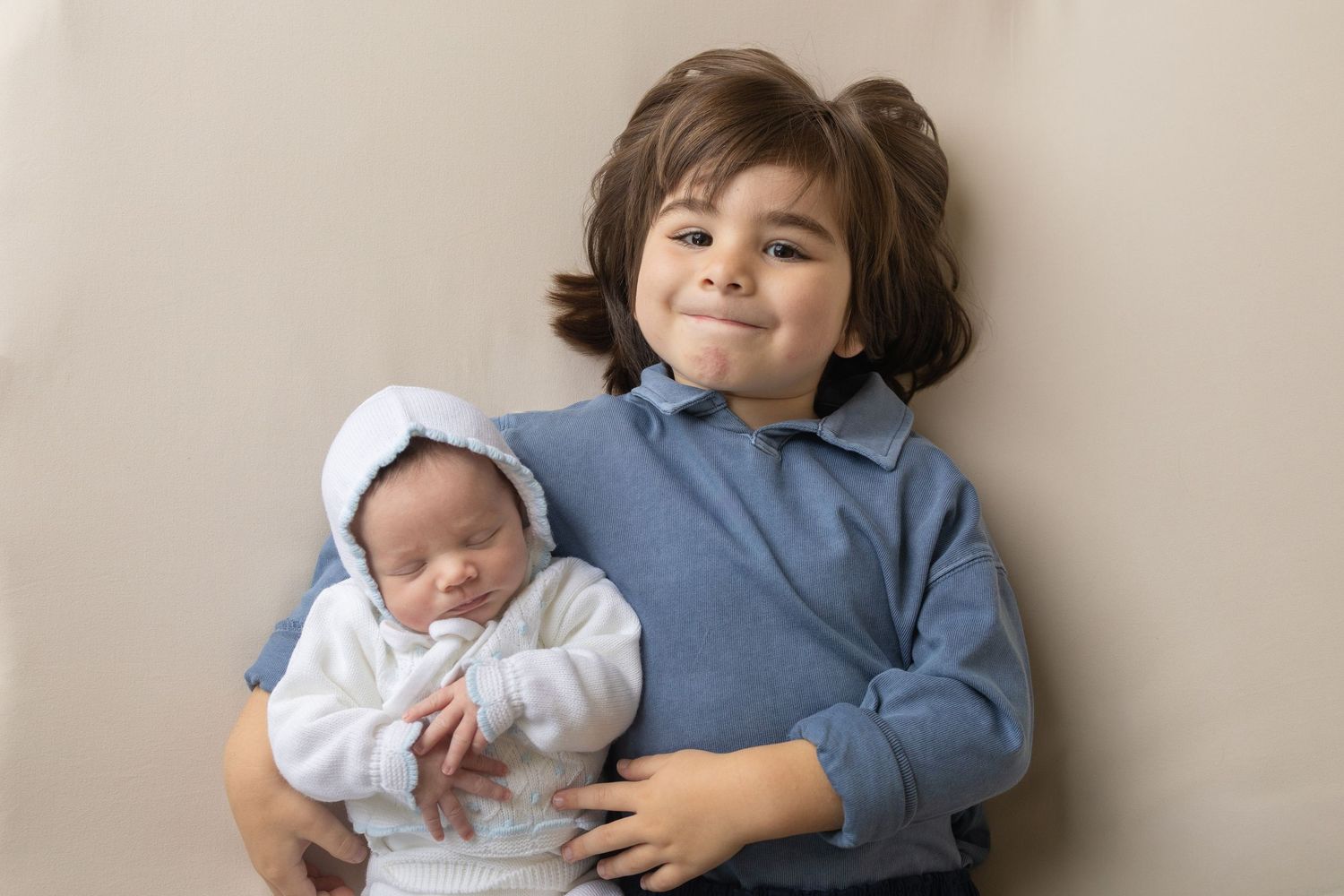 Young boy smiling and gently holding a sleeping newborn baby dressed in white, posed against a neutral background.