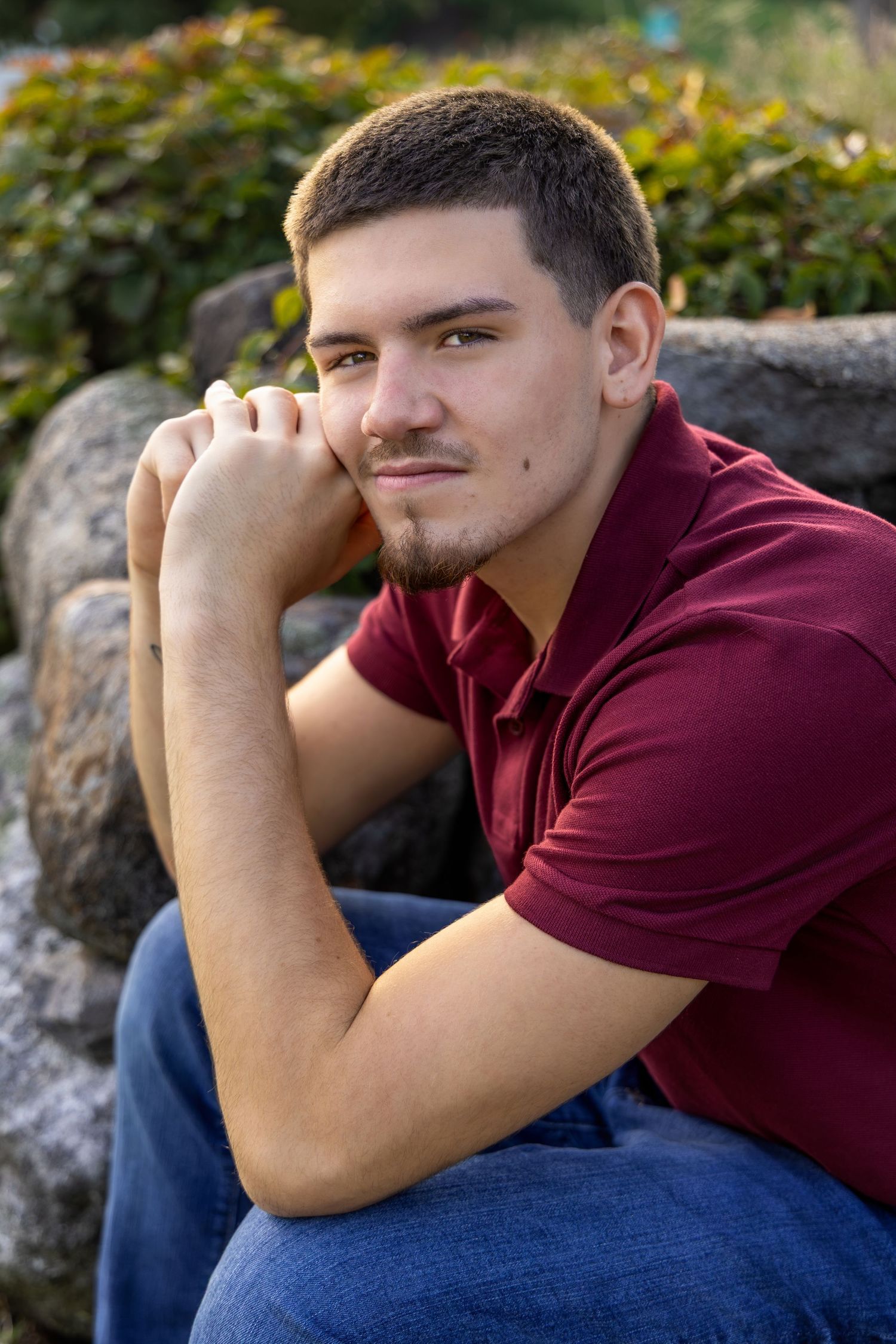 Young man in maroon polo shirt sitting outdoors on rocks, resting chin on hands, with greenery background.