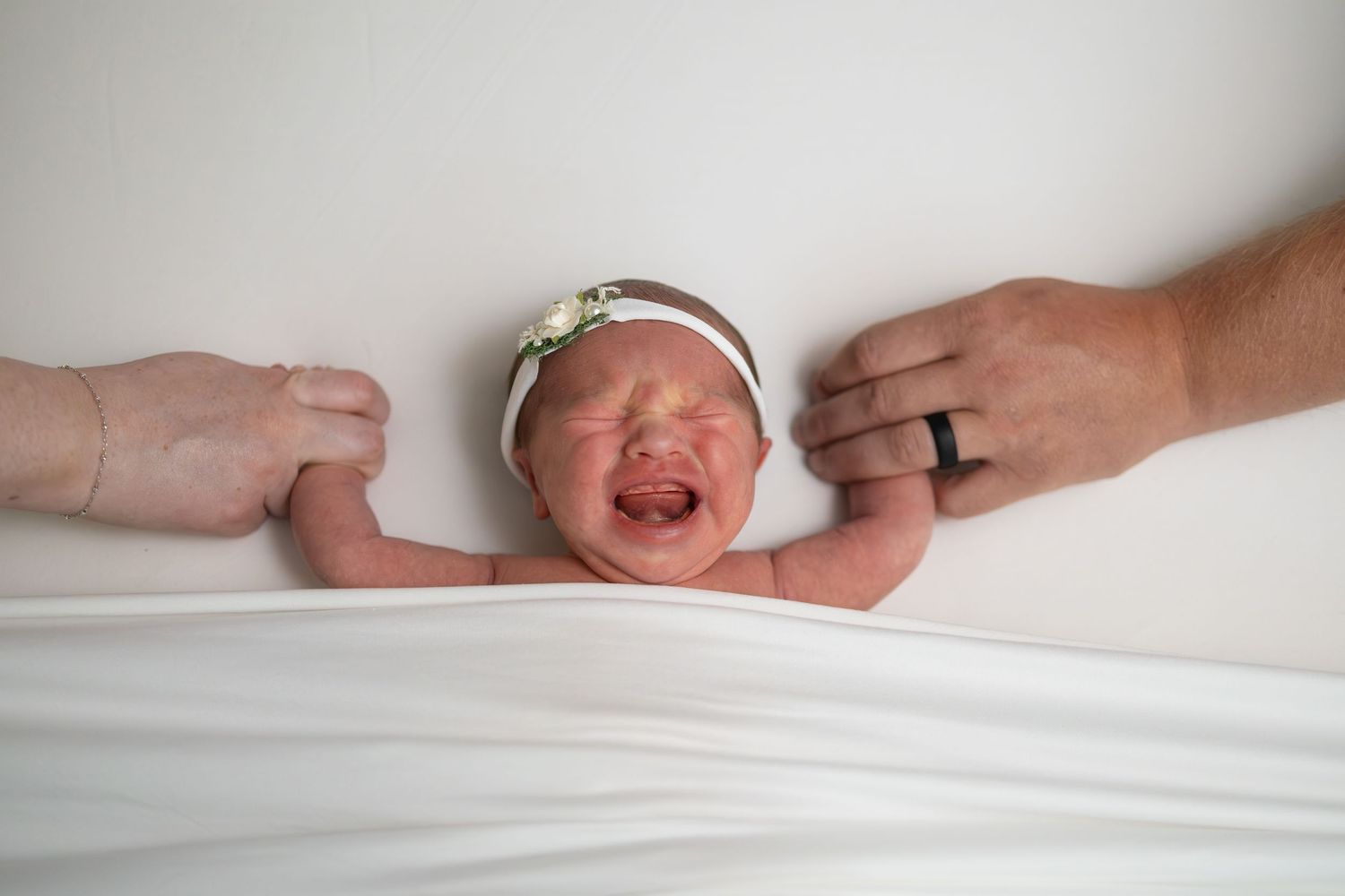 Crying newborn baby girl wearing a floral headband, held by two adults on a white blanket.