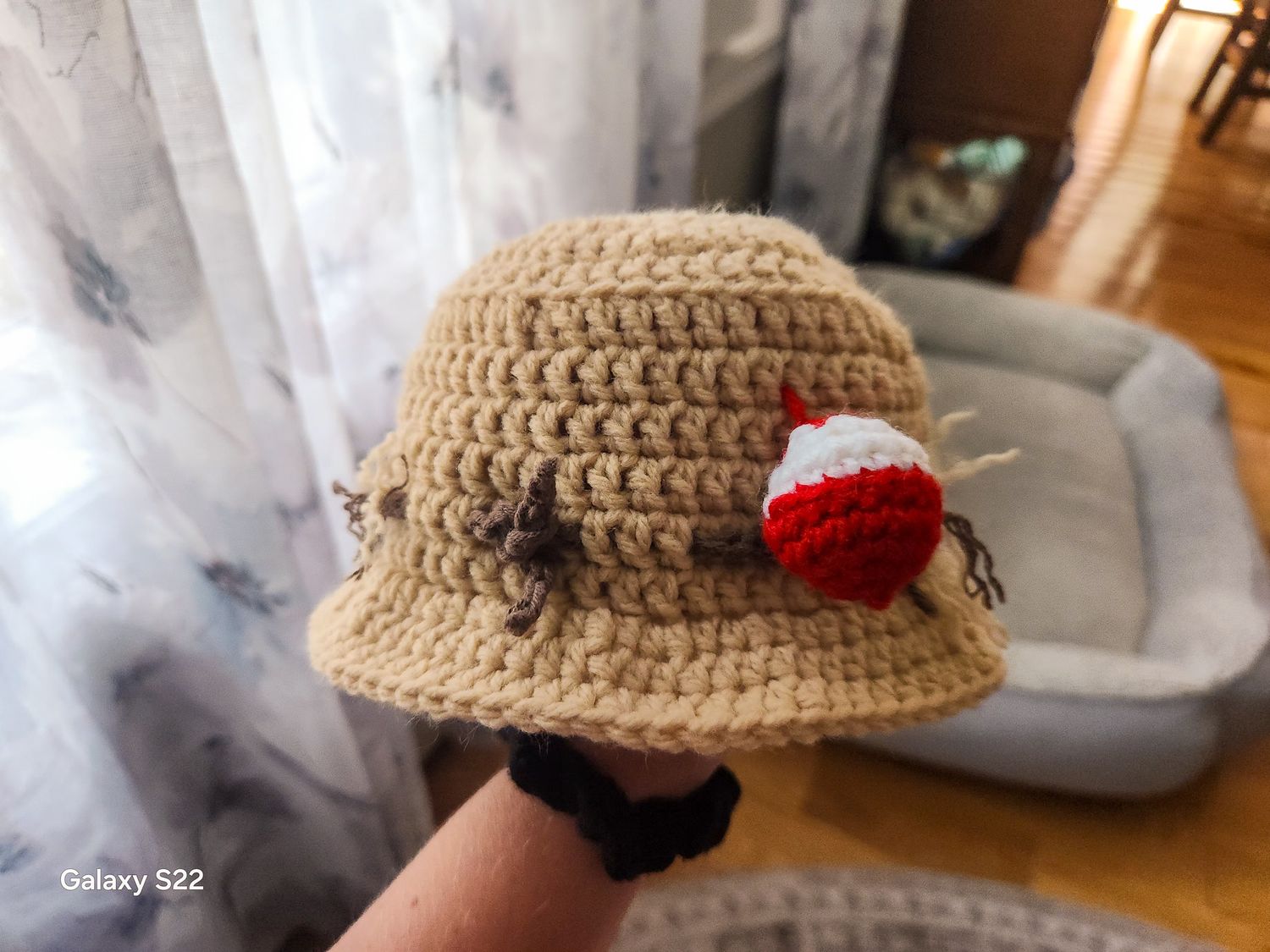 Handmade crochet bucket hat in tan yarn with fishing bobber and fly decorations displayed outdoors.