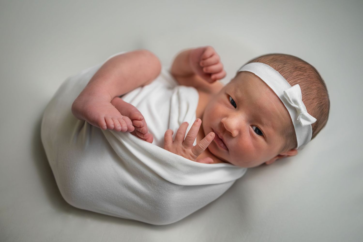 Newborn baby swaddled in white wrap with bow headband, peacefully resting on soft white background.