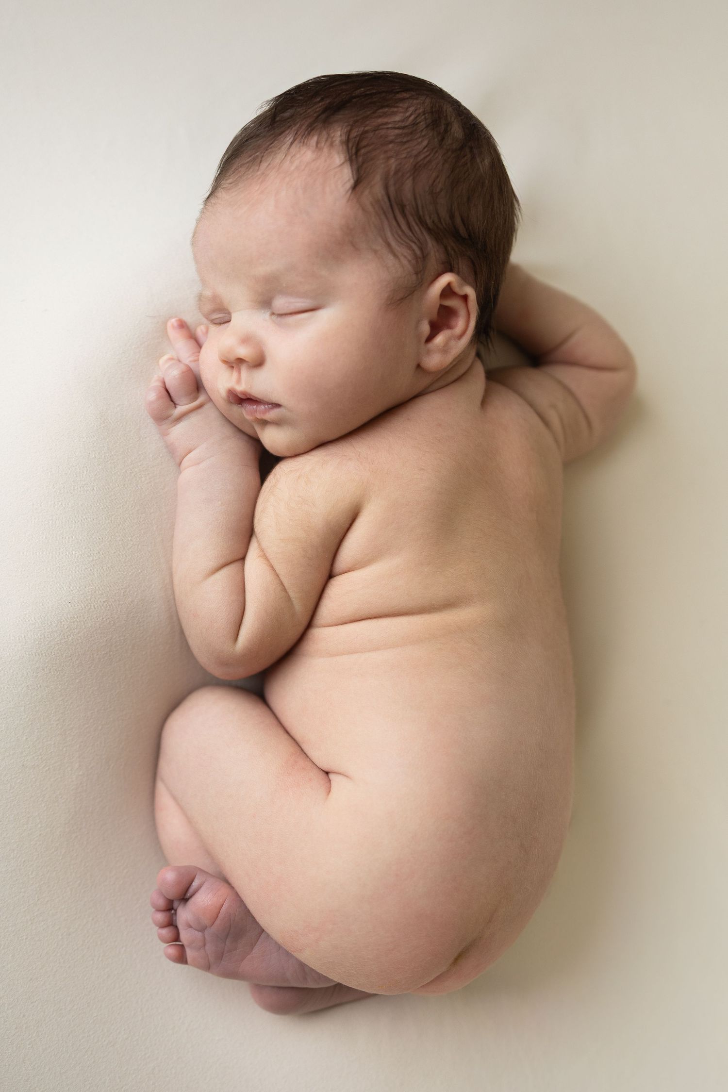 Peaceful newborn baby sleeping curled up on a soft cream blanket in a classic newborn photography pose.