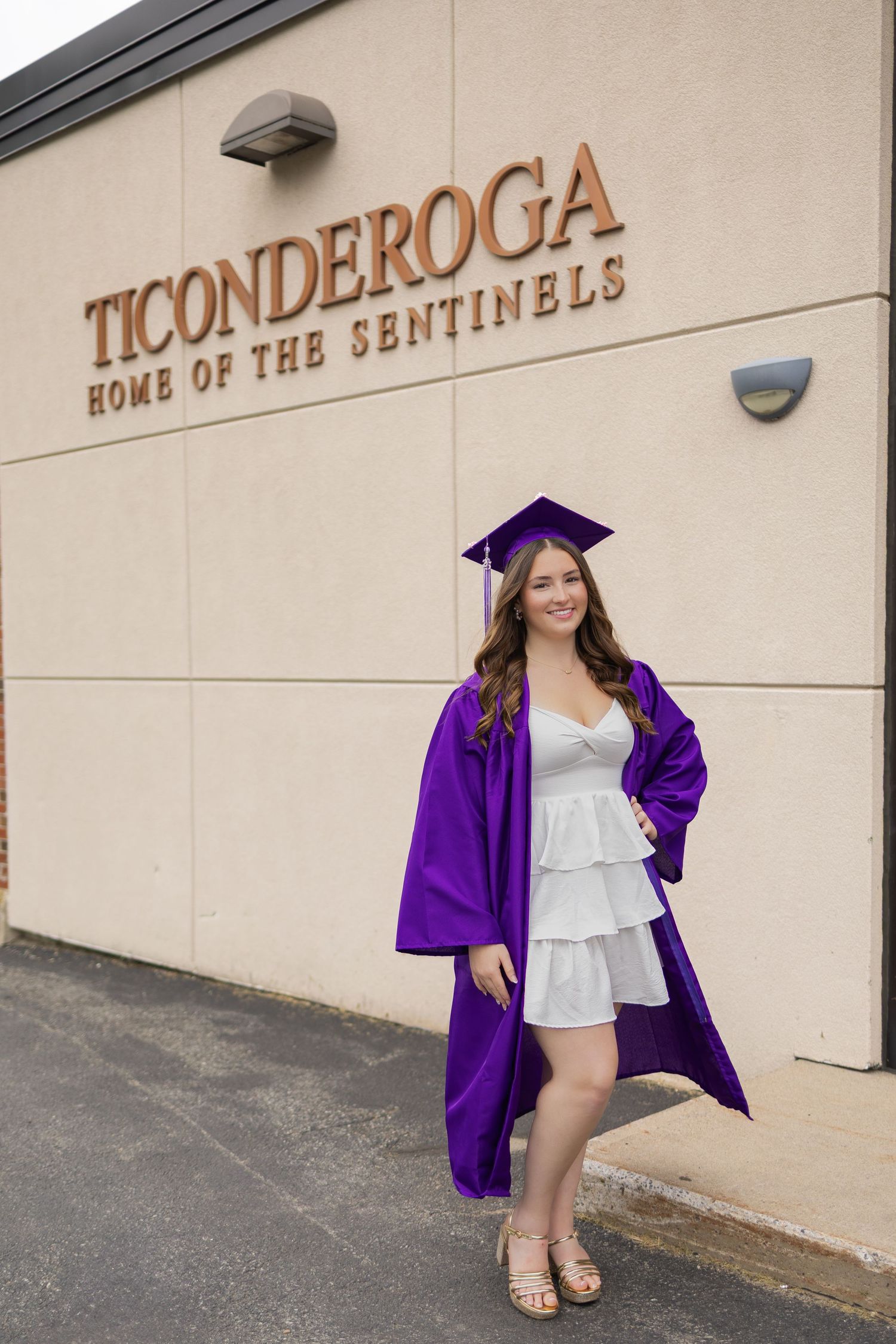 Graduate in purple cap and gown poses against beige wall with Ticonderoga signage.