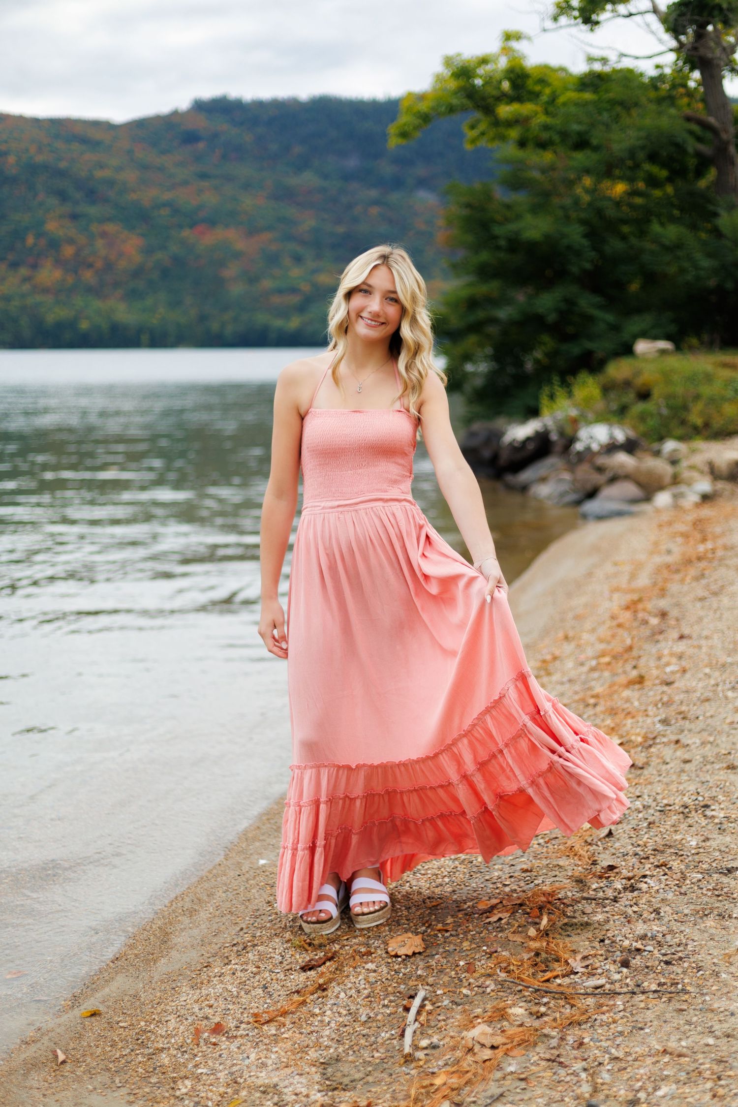 A coral pink maxi dress flows gracefully along a rocky lakeside beach with mountains in the background.