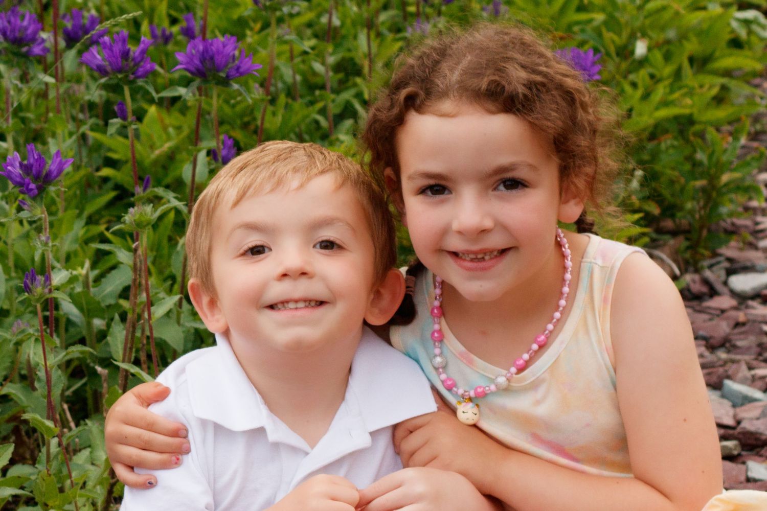 Children smiling together among purple flowers in a sunny garden setting.