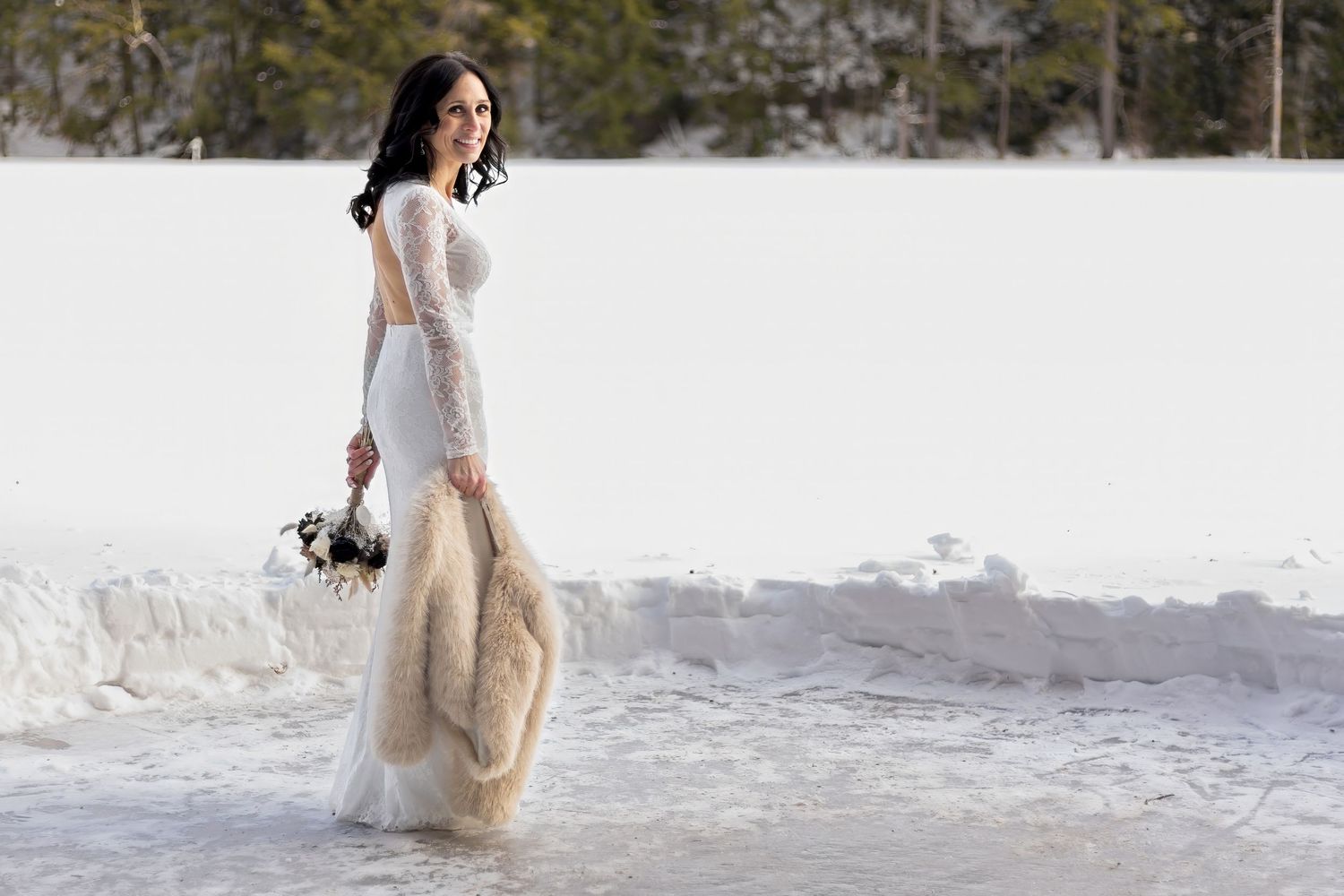 Bride in white dress and fur shawl walking through snowy landscape.