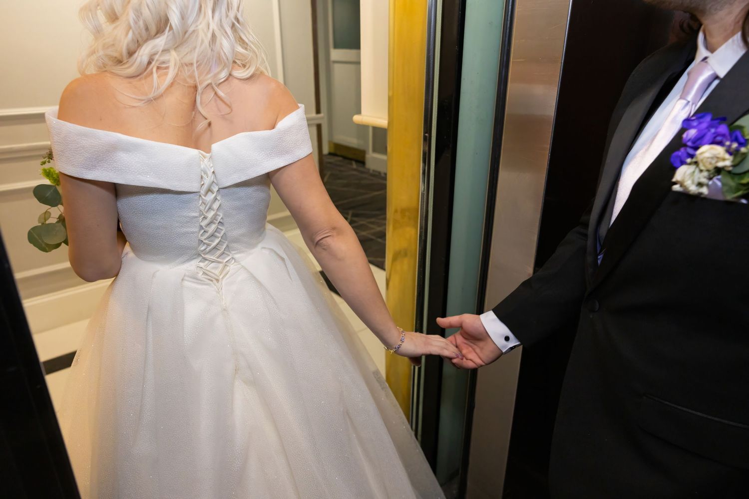 A couple stands together in a hallway, the bride wearing an elegant off-shoulder white gown.