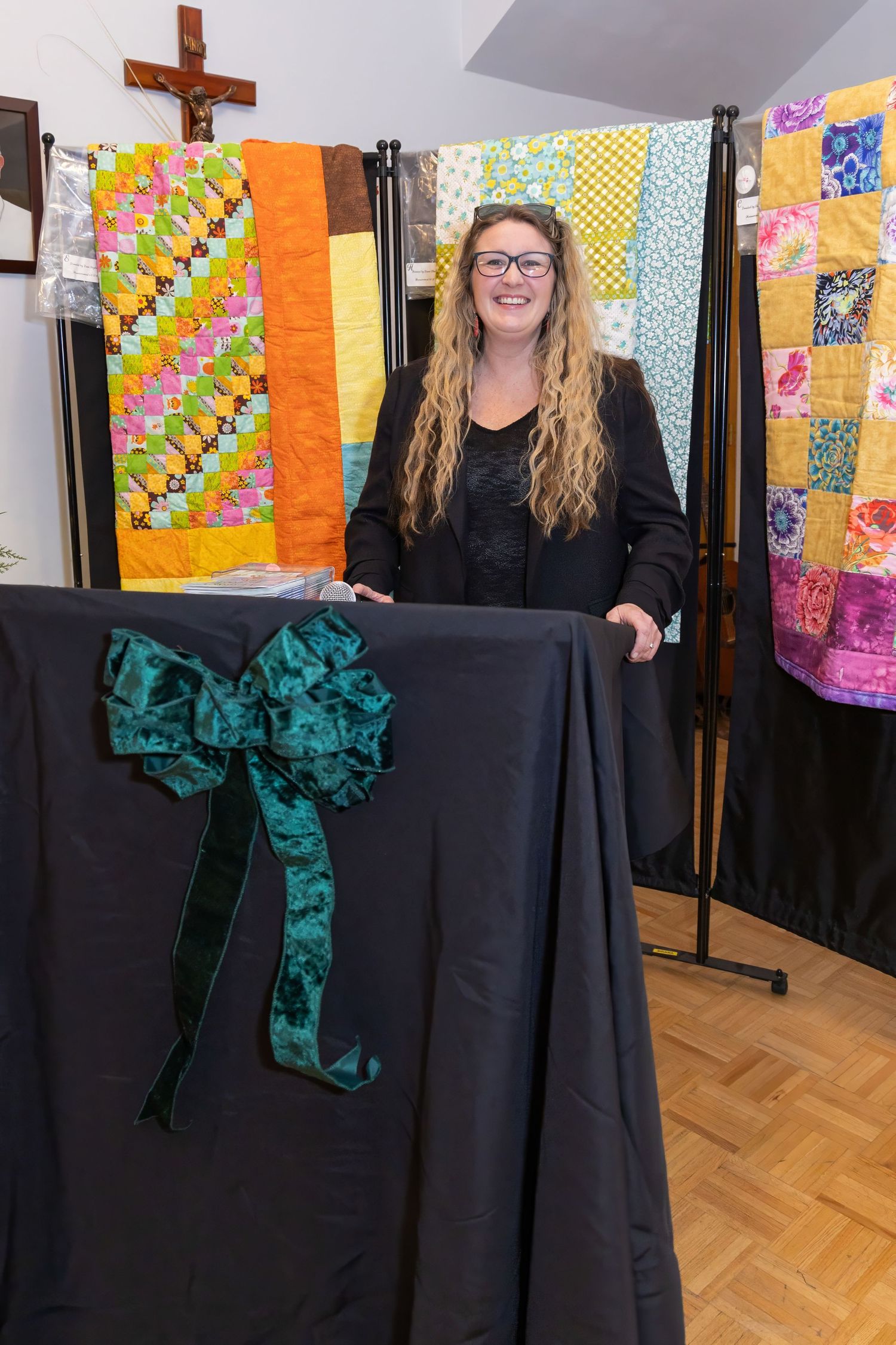 A performer in a black dress with emerald green accents stands in front of colorful quilt displays.