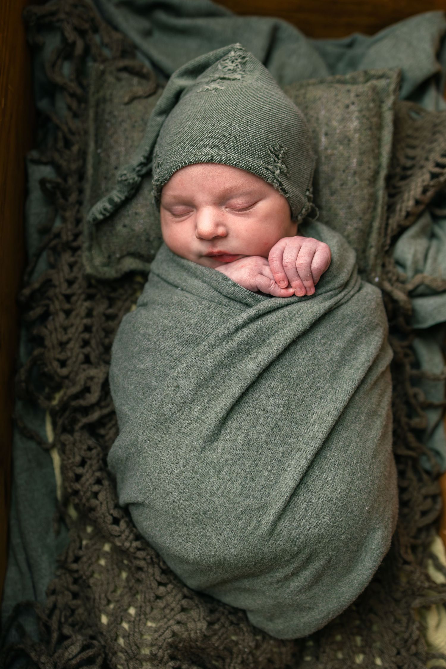 Sleeping newborn baby swaddled in a green wrap and matching knit hat, resting peacefully in a wooden box.