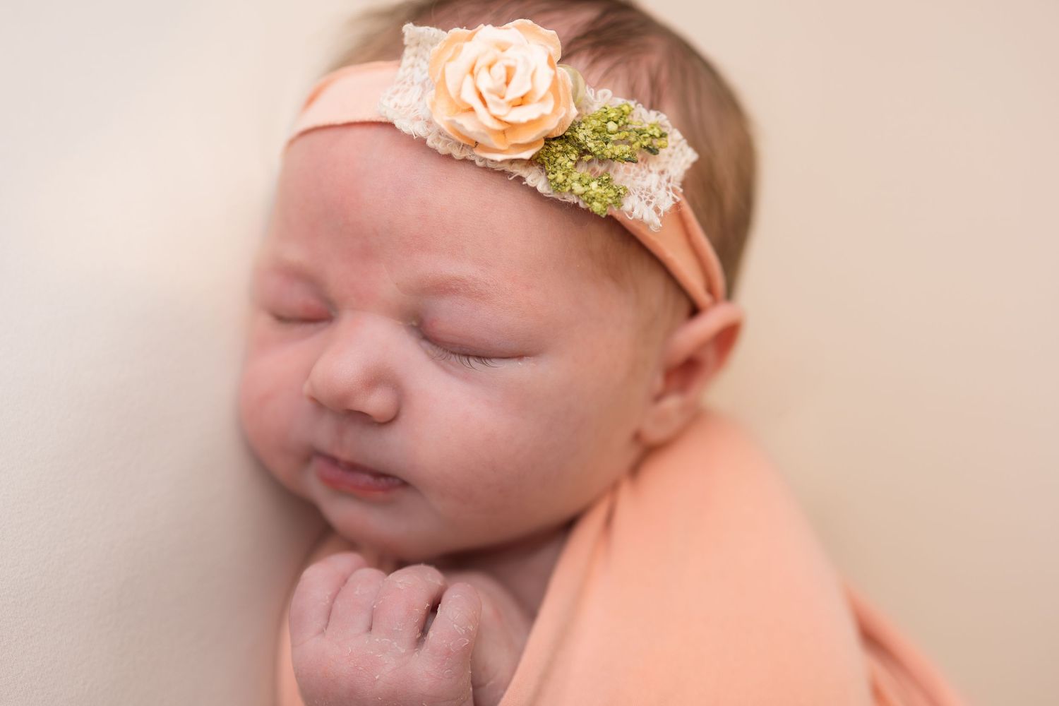 Peaceful newborn baby girl sleeping wrapped in peach fabric wearing a floral headband with rose and green accents.