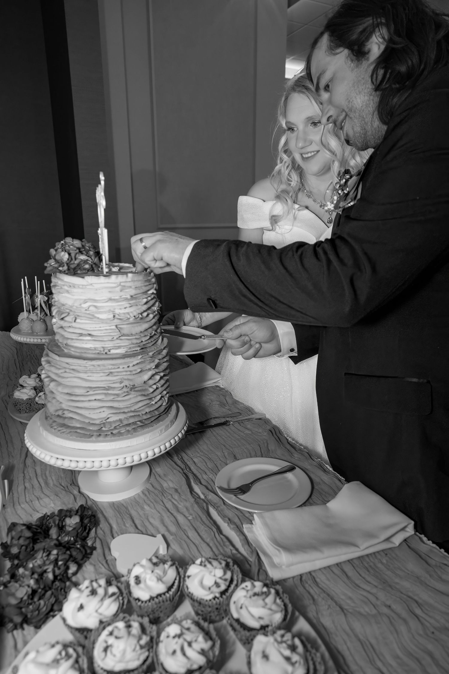 Black and white photo of wedding cake cutting with cupcakes displayed on table.