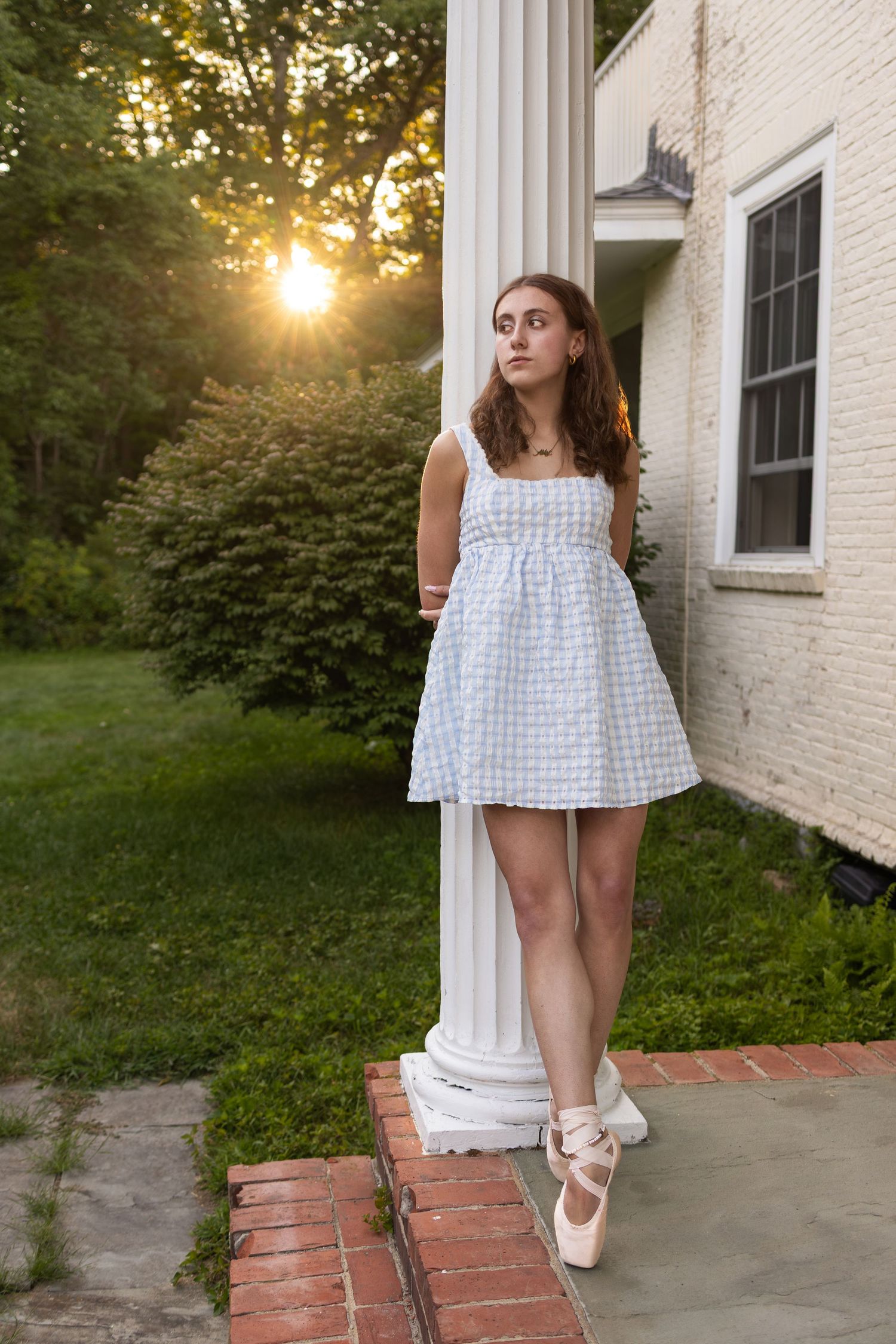 Young woman in blue gingham mini dress leaning against white column at sunset near brick house.