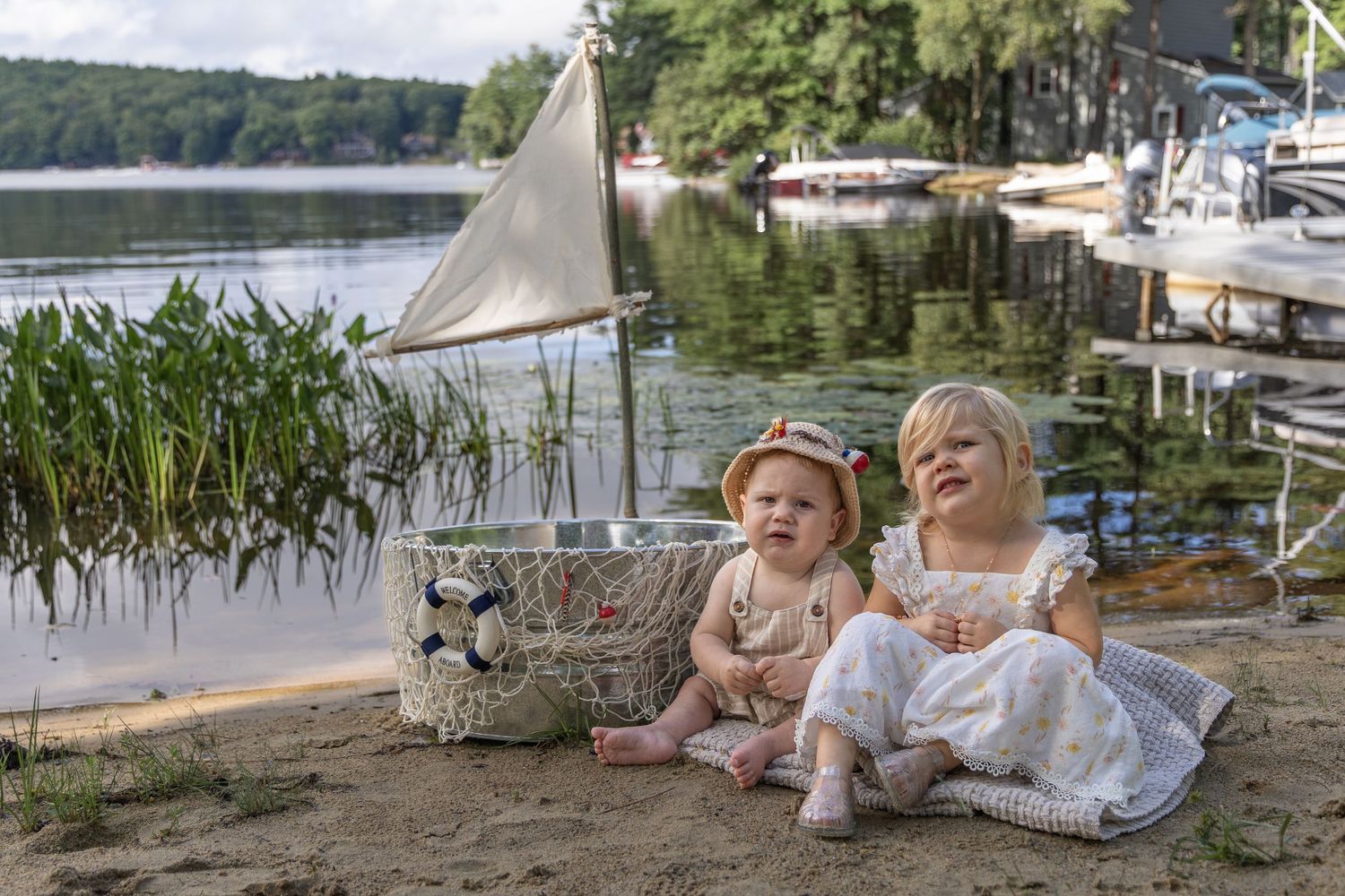 Two young children sit on a sandy lakeside beach beside a decorative basket with a sail, boats visible in the background.