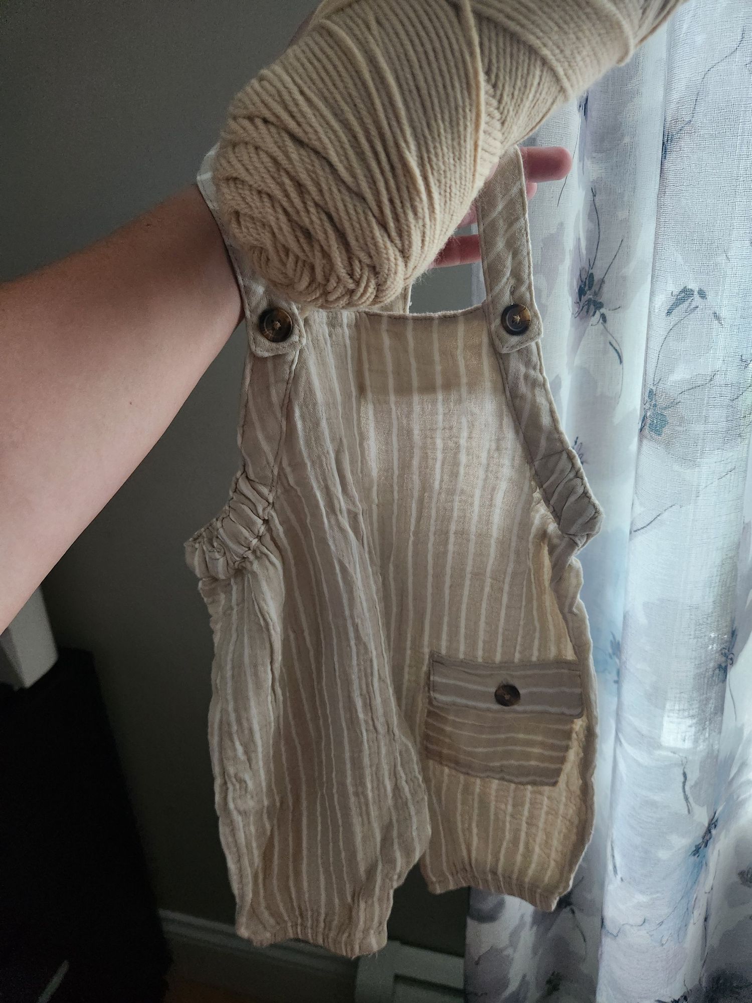 Hand holding tiny striped linen baby romper overalls with button details and front pocket.
