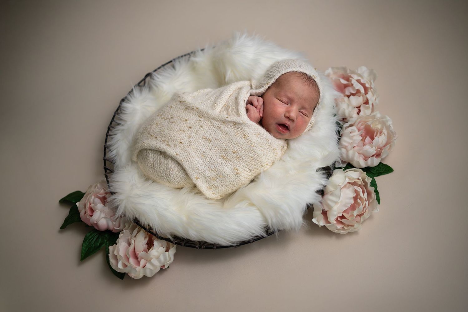 Newborn baby sleeping peacefully in a bowl with white fur and pink peonies, wrapped in a cream knit swaddle.