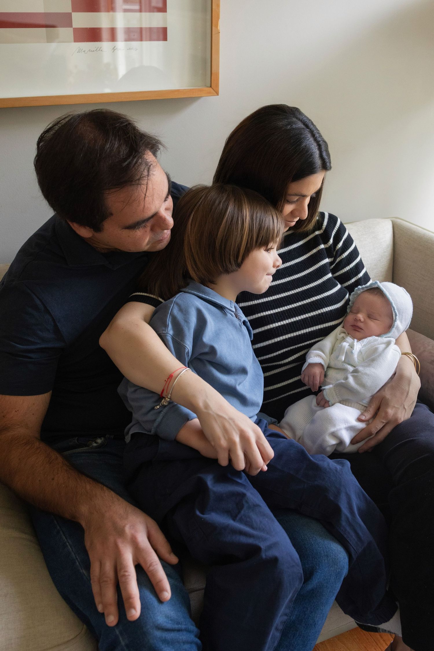 Family of four sitting on a couch, parents admiring their newborn baby while older child looks on.