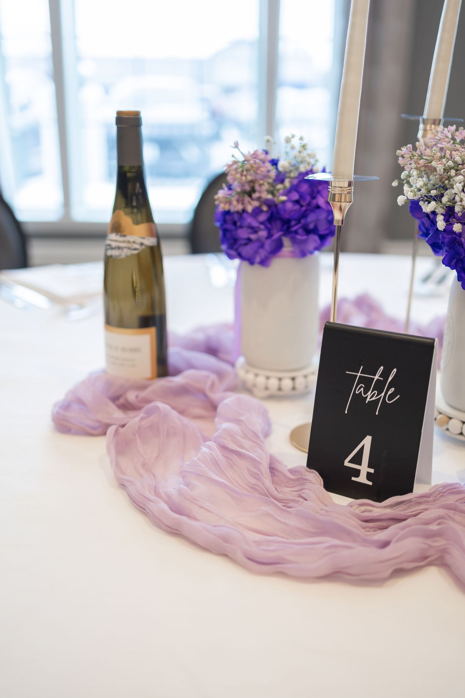 Wedding table decor with purple flowers, wine bottles and table numbers on lavender fabric.