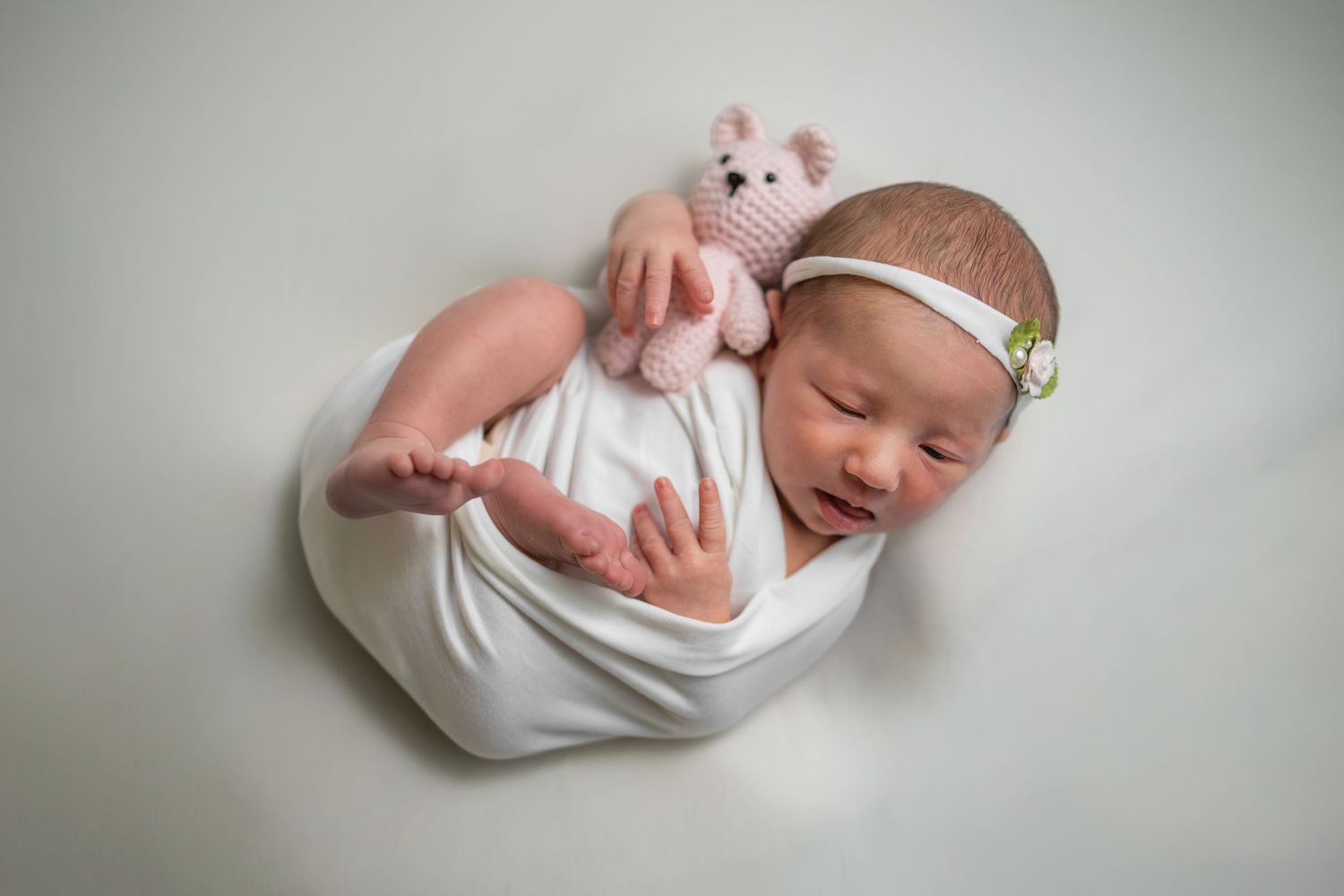 Sleeping newborn baby wrapped in white swaddle holding a pink crocheted teddy bear with a floral headband.