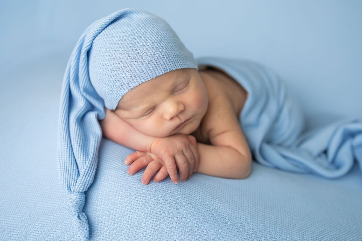 Peaceful newborn baby sleeping in a blue knit hat and wrap on a soft blue background.