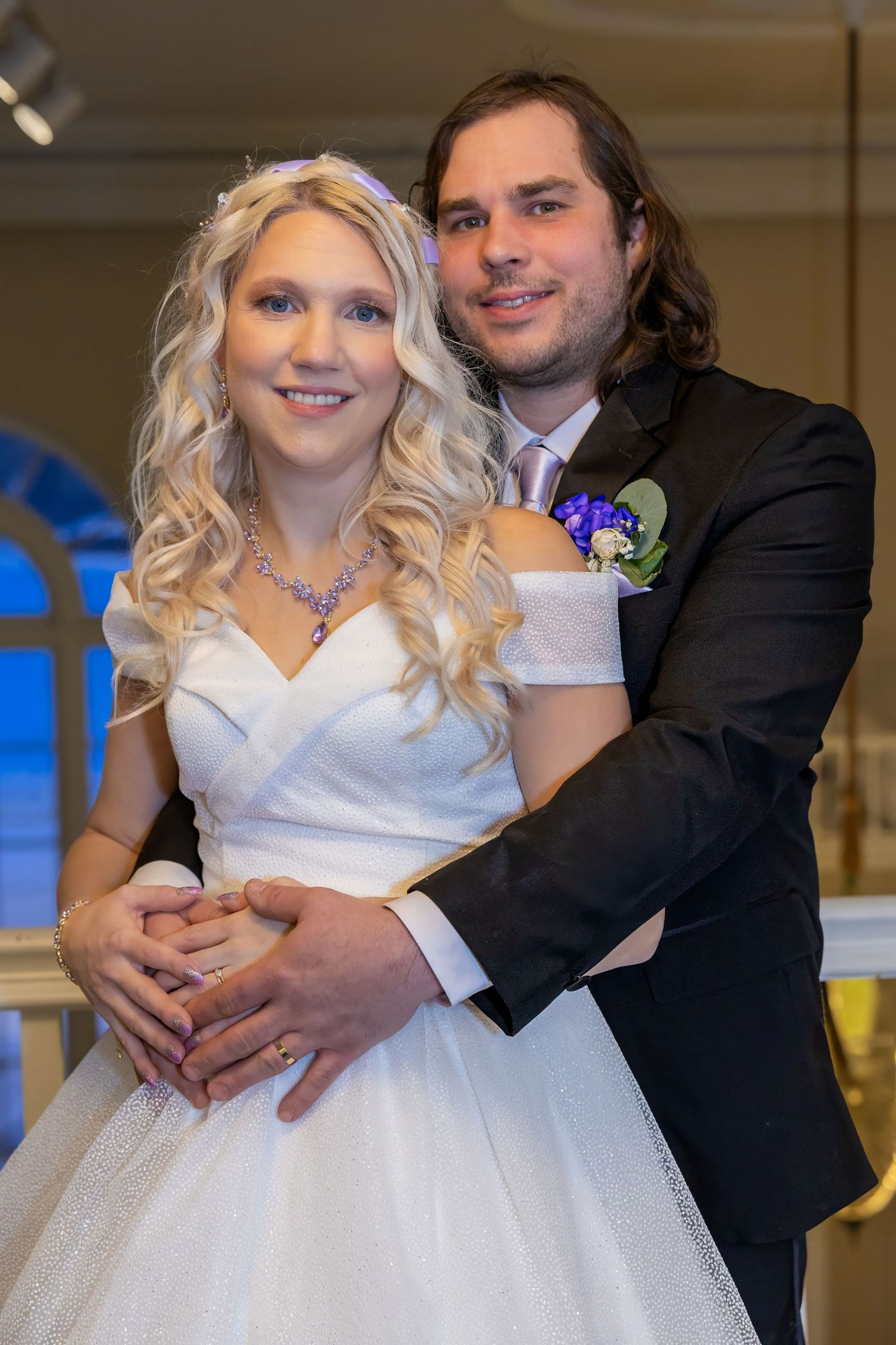 A couple embraces in a room with large windows during their wedding celebration.