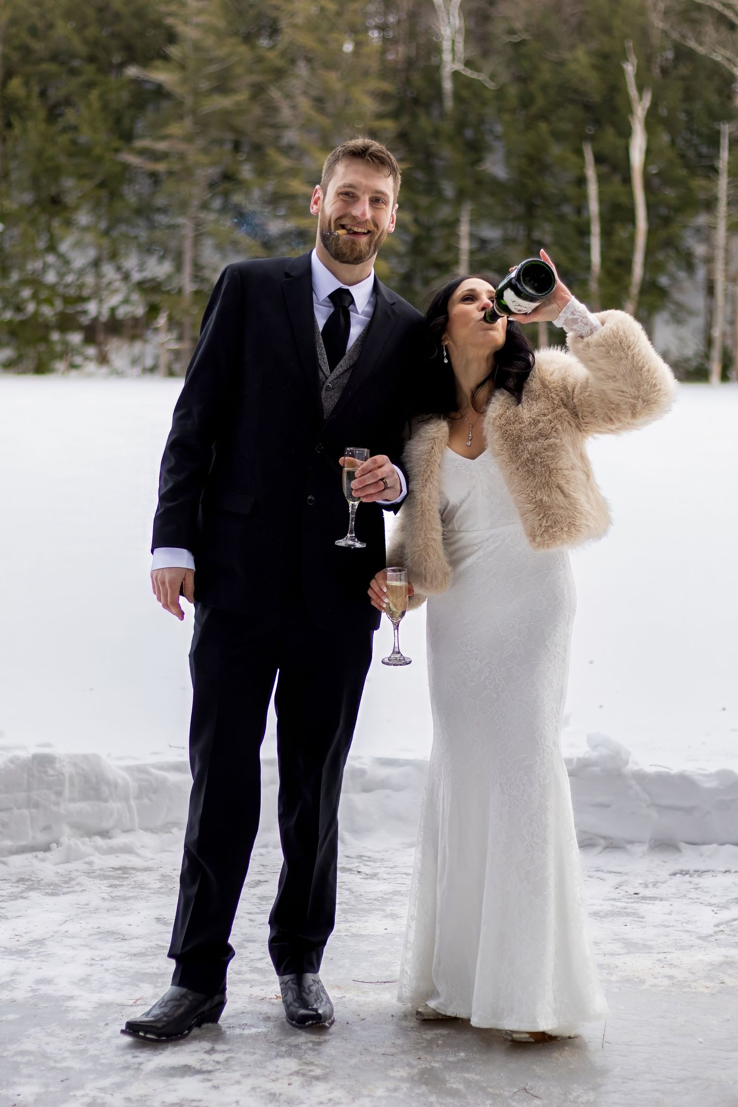 Wedding couple sharing champagne and laughing in snowy winter setting.