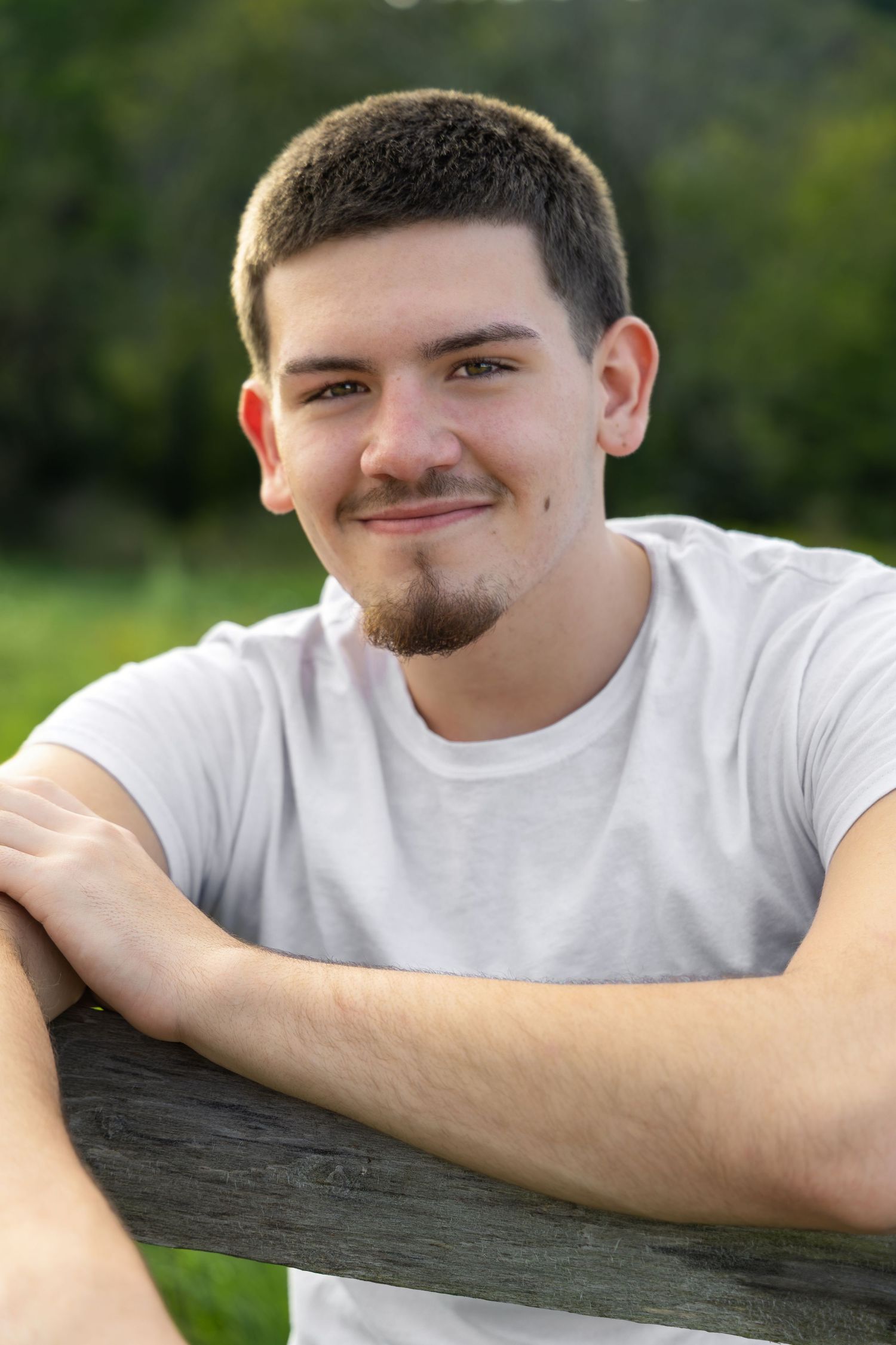 Young man with short hair and goatee smiling outdoors, leaning on wooden fence in white t-shirt.