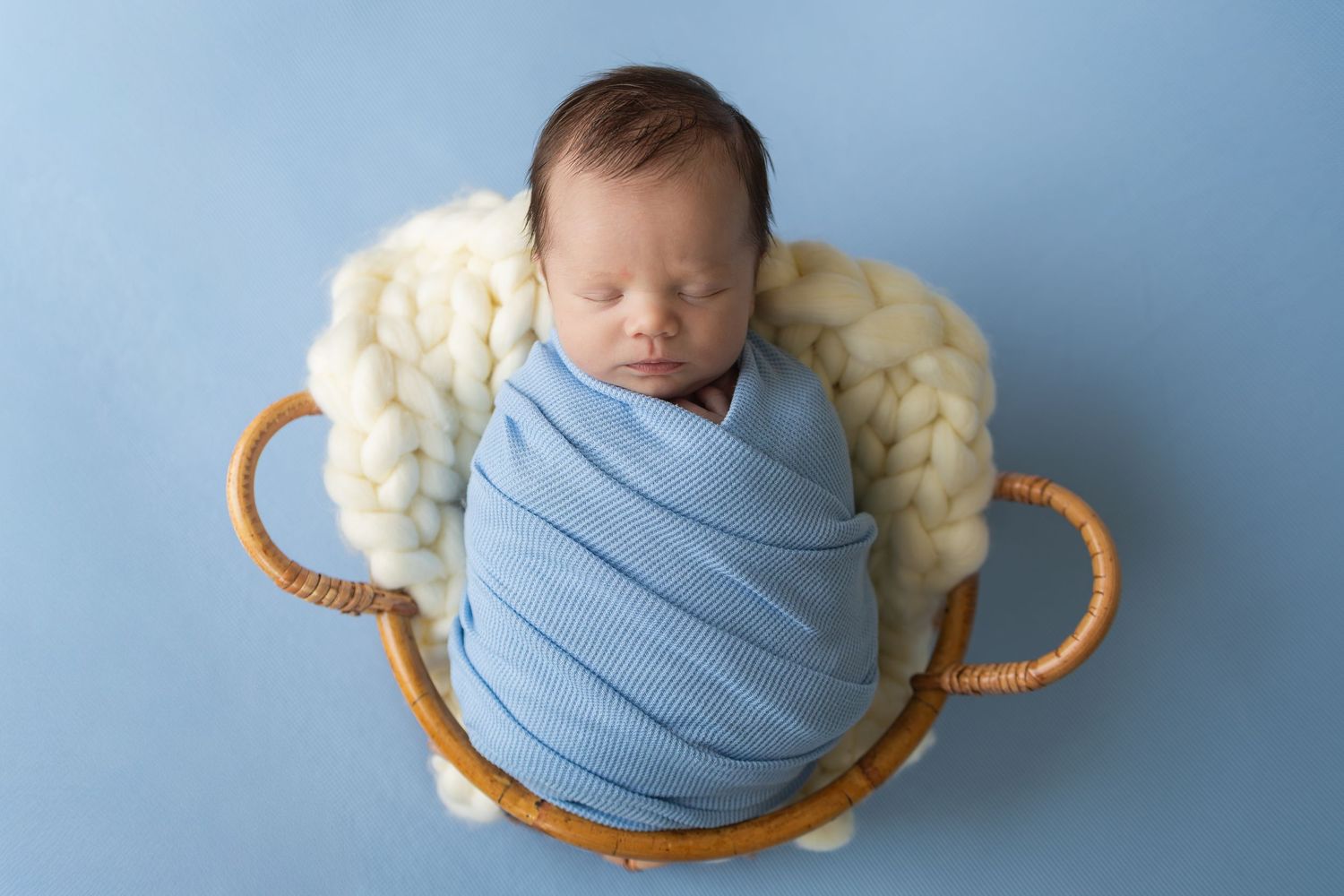 Sleeping newborn baby swaddled in blue wrap nestled in a wicker basket with white wool on a blue background.