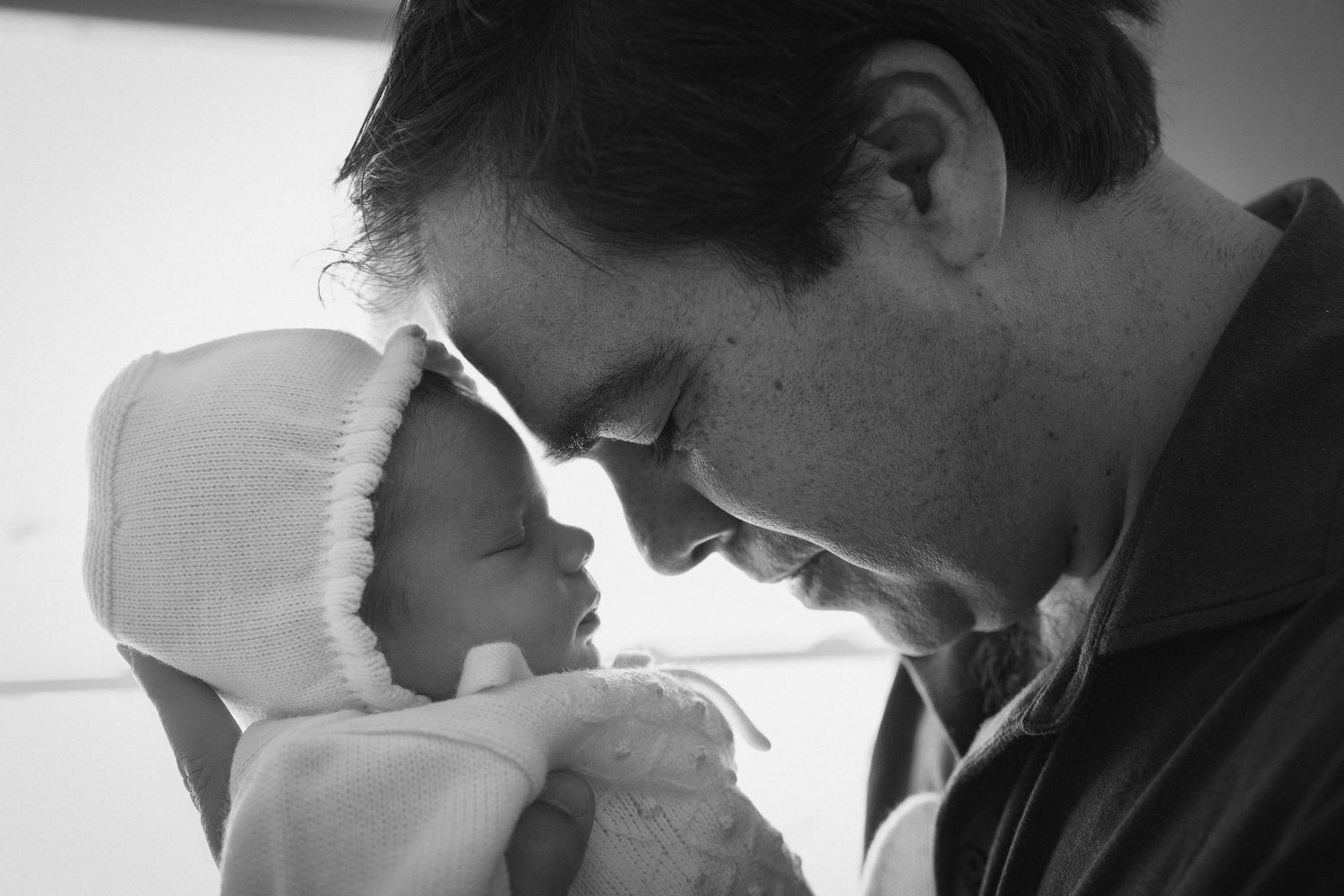 Black and white photo of a father tenderly touching foreheads with his newborn baby wrapped in a white blanket.