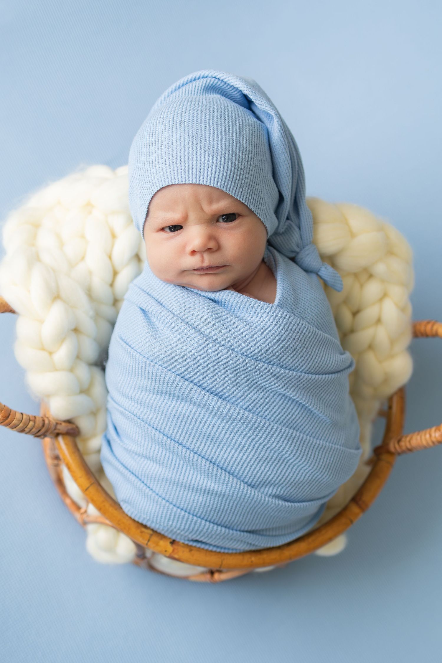 Newborn baby swaddled in blue wrap and hat, resting in a wicker basket with white chunky knit blanket.