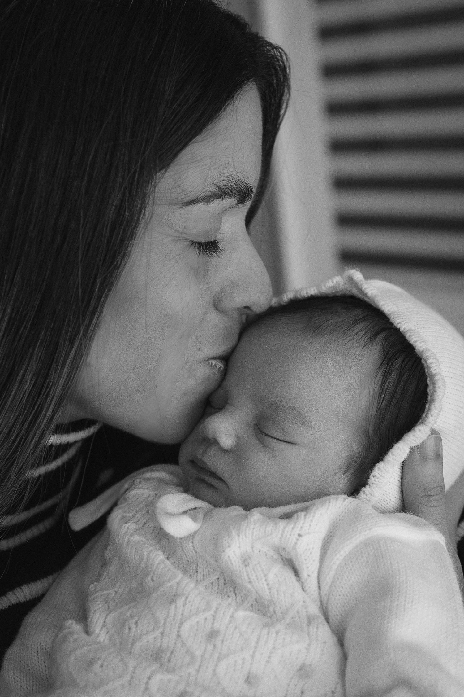 Black and white photo of a mother gently kissing her sleeping newborn baby wrapped in a white knit outfit.