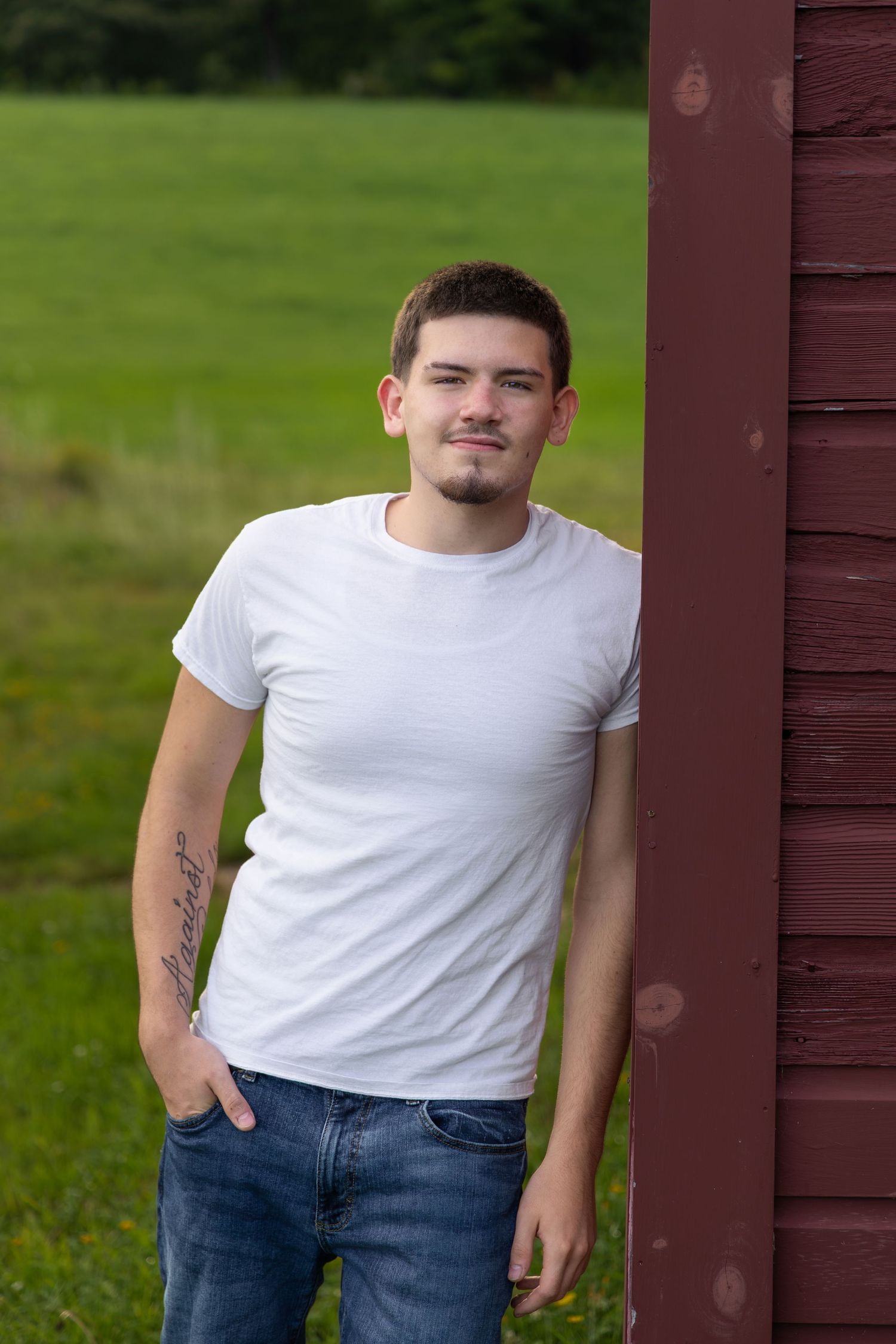 Young man with tattoo in white t-shirt and jeans leaning against a red barn with green field background.