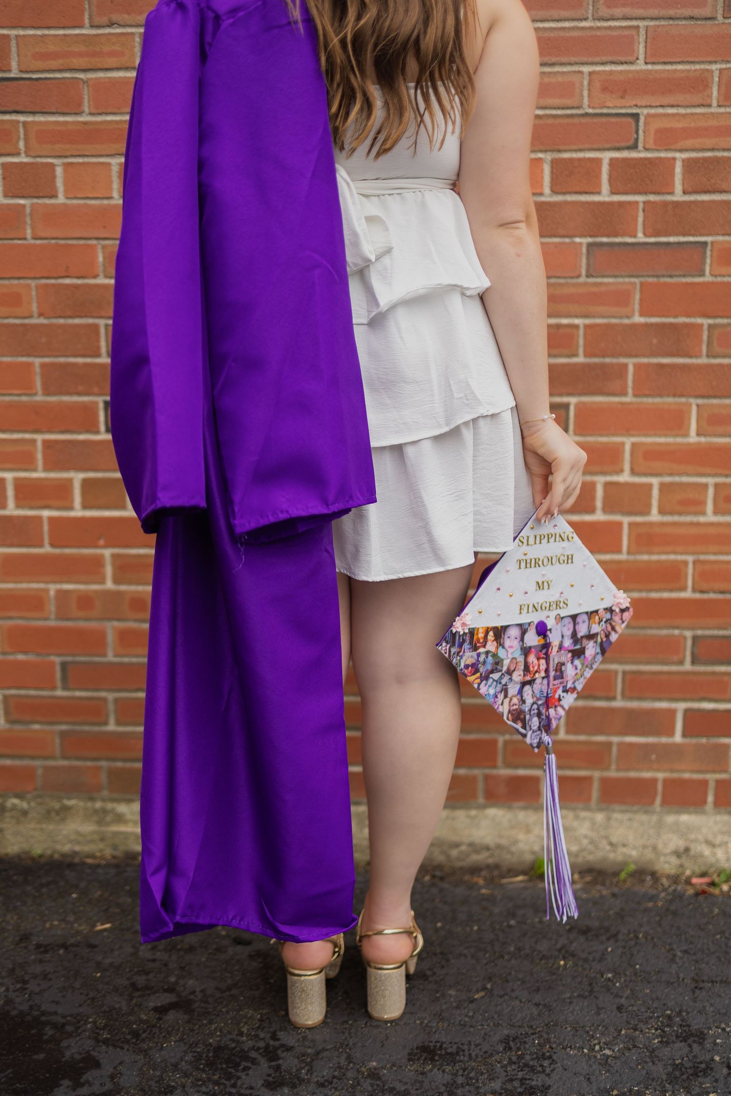 Graduate poses with decorated cap against brick wall in white dress and purple gown.