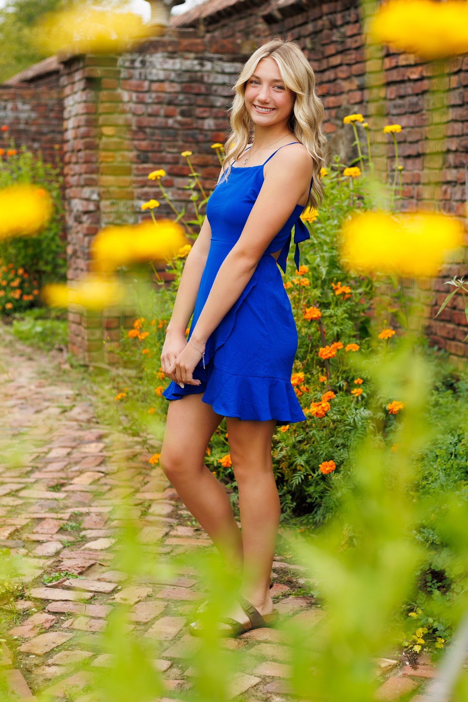 Vibrant garden portraits with yellow flowers and brick wall backdrop featuring a blue dress.