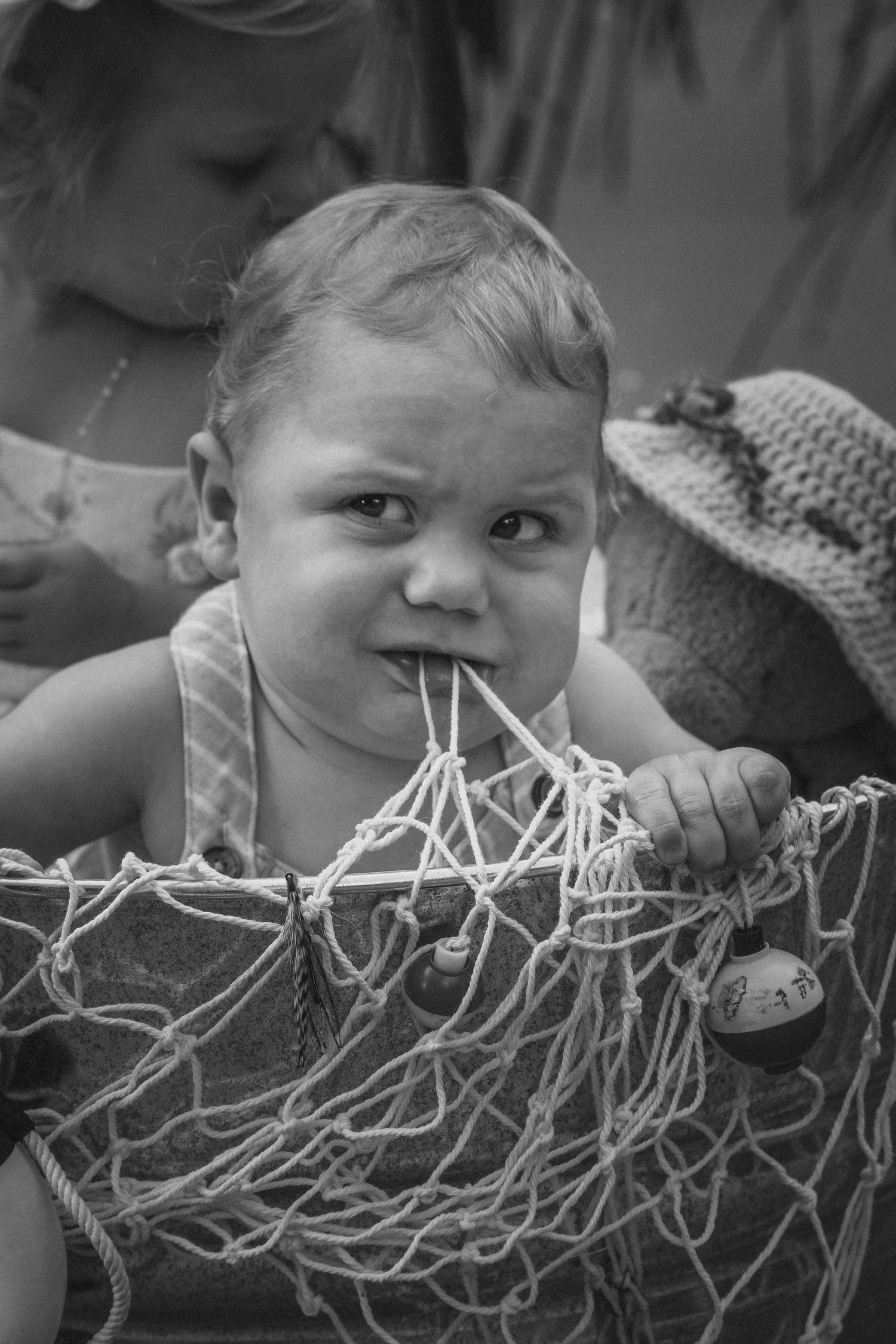Black and white photo of a curious baby chewing on a fishing net, held by an adult in the background.