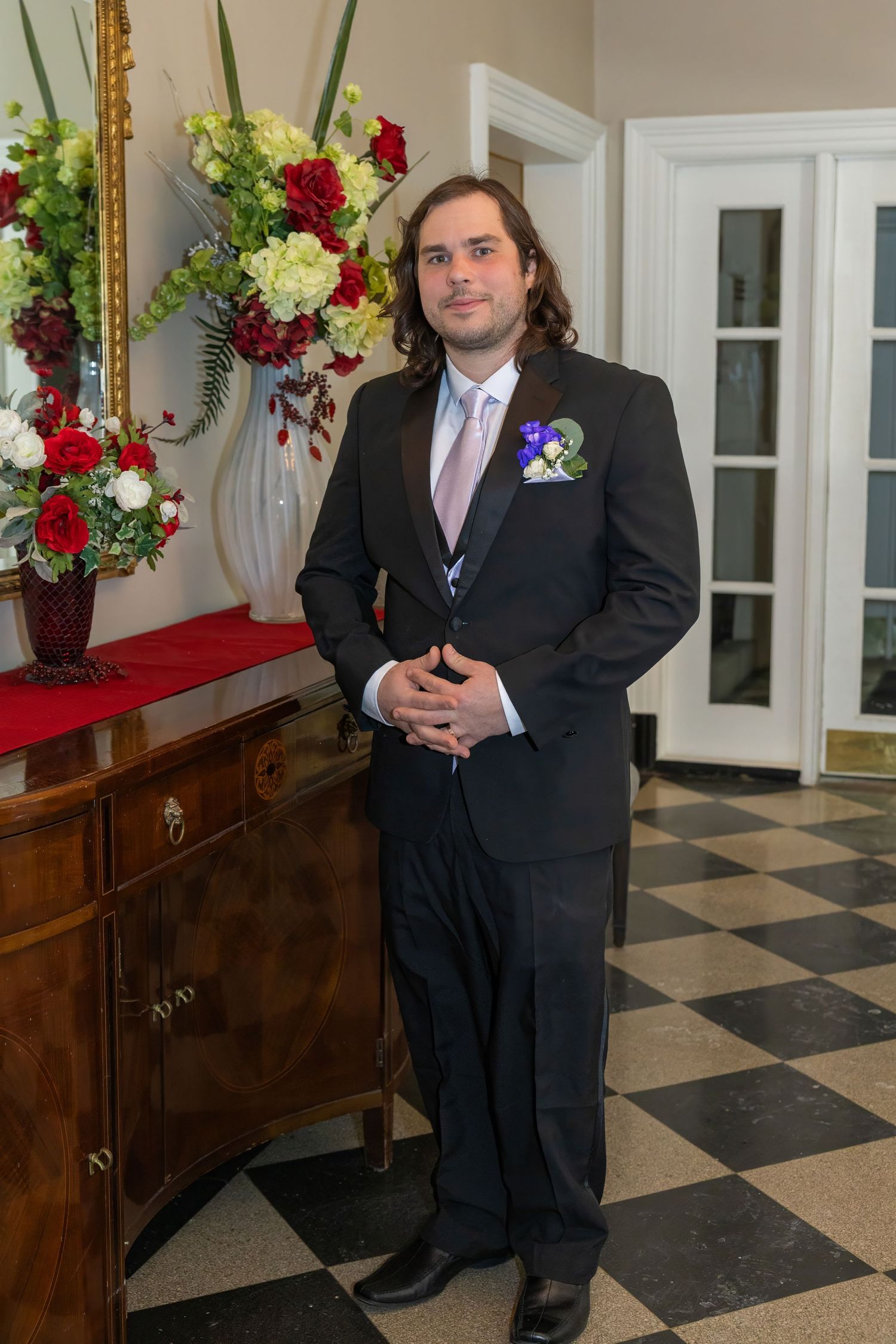 A person in a black tuxedo stands in a hallway with checkered floors and elegant floral arrangements.