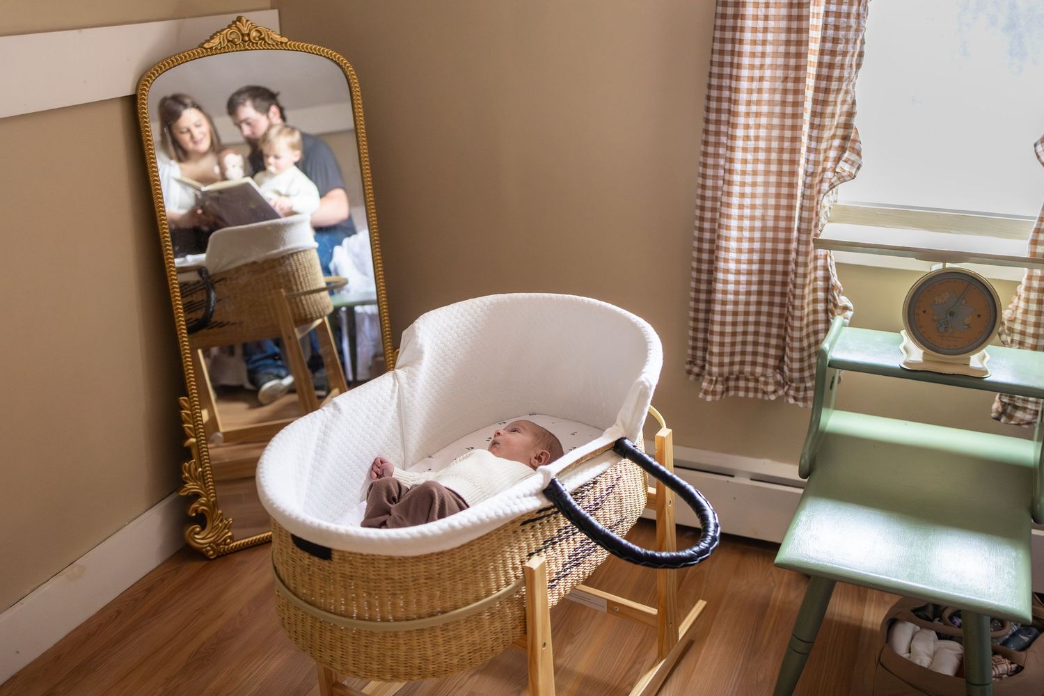 Newborn baby sleeping in a wicker Moses basket with parents reflected in a gold ornate mirror nearby.