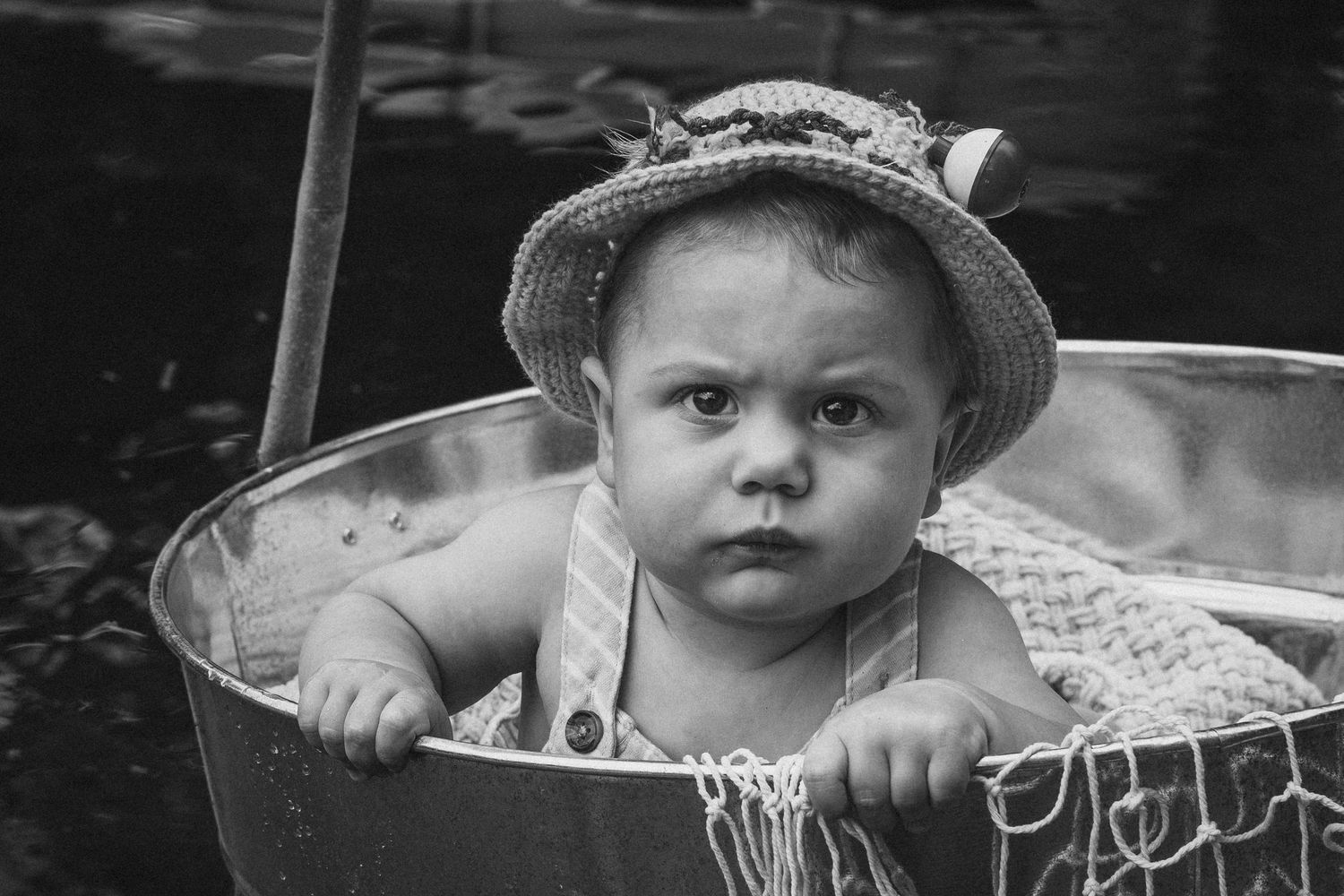 Black and white photo of a cute baby wearing a straw hat sitting in a metal tub outdoors.