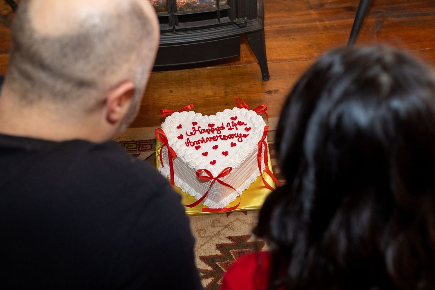 A couple looks down at a heart-shaped cake with red decorations near a fireplace.