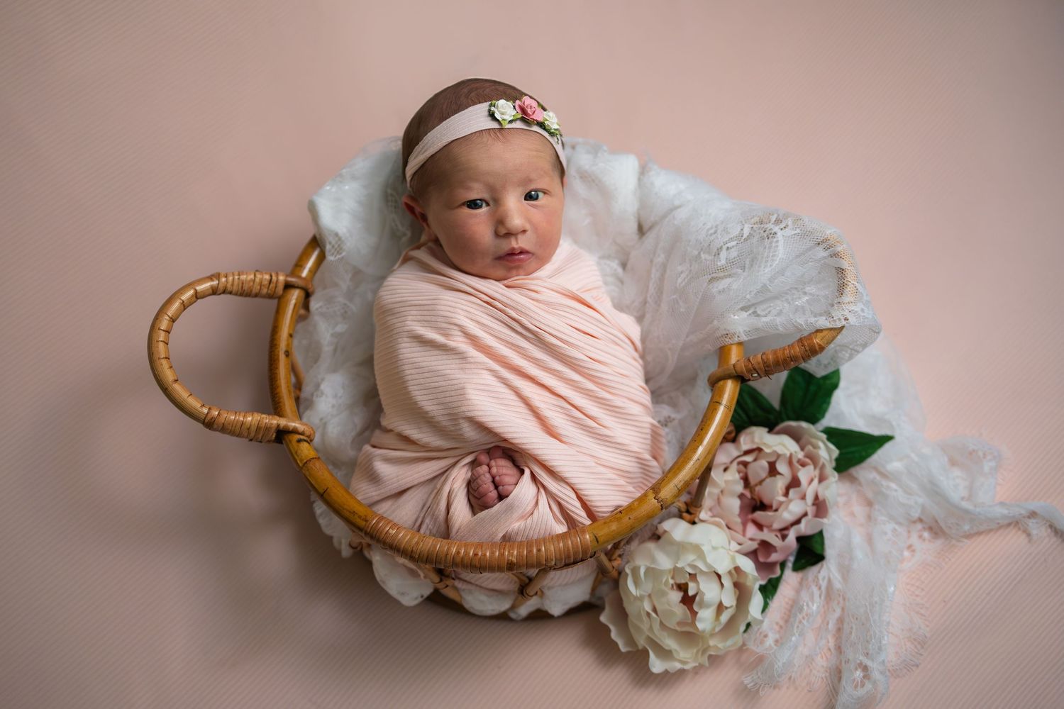 Newborn baby girl swaddled in pink wrap, resting in a wicker basket with white lace and peony flowers.
