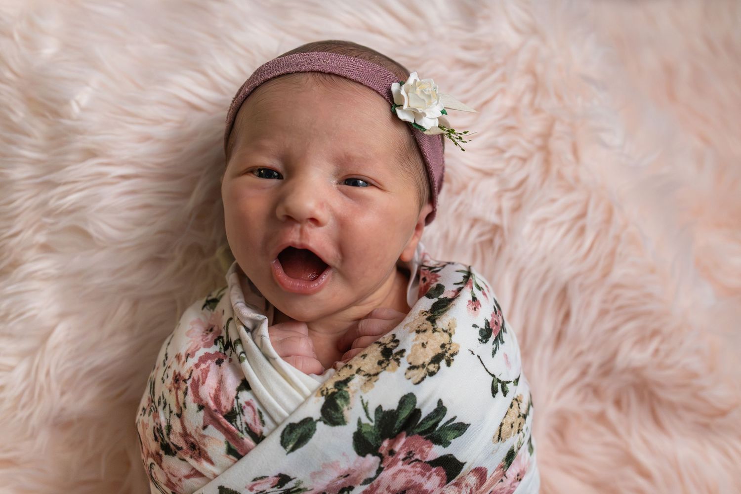 Newborn baby girl swaddled in floral wrap with pink headband, smiling on fluffy pink fur blanket.