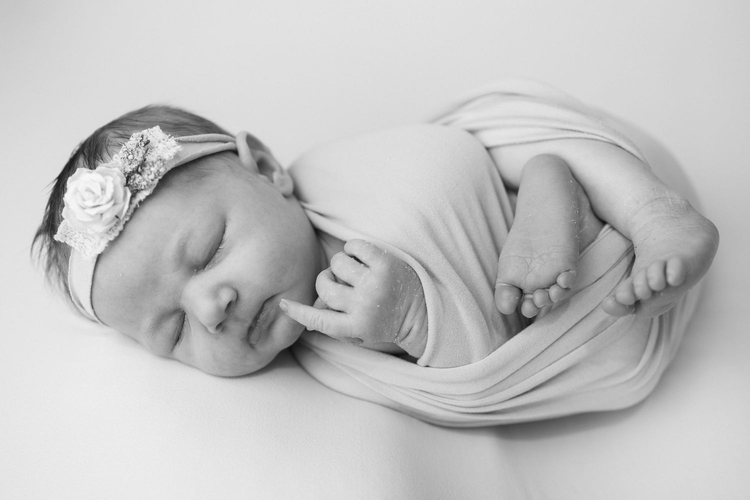 Black and white newborn photo of a sleeping baby girl wrapped in soft fabric wearing a floral headband.