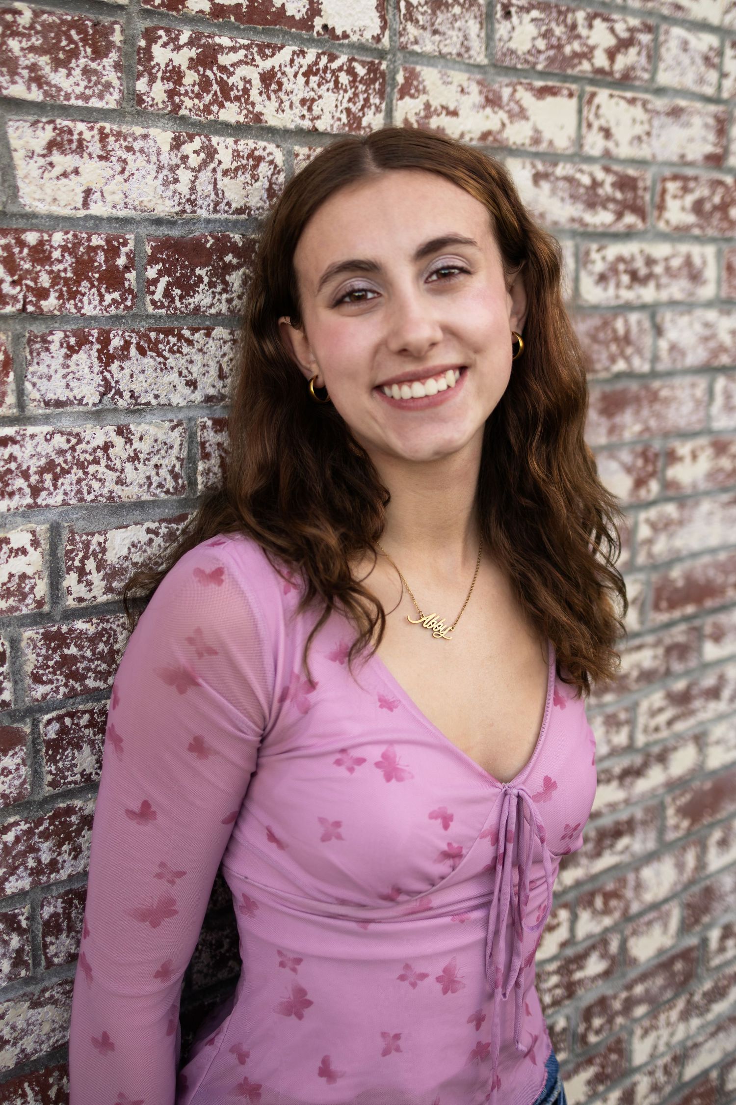 Smiling young woman in pink floral top leaning against rustic brick wall wearing gold necklace.